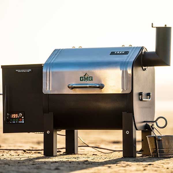 Blue and black GMG pellet smoker on a sandy outdoor surface, with chimney and smoke stack.