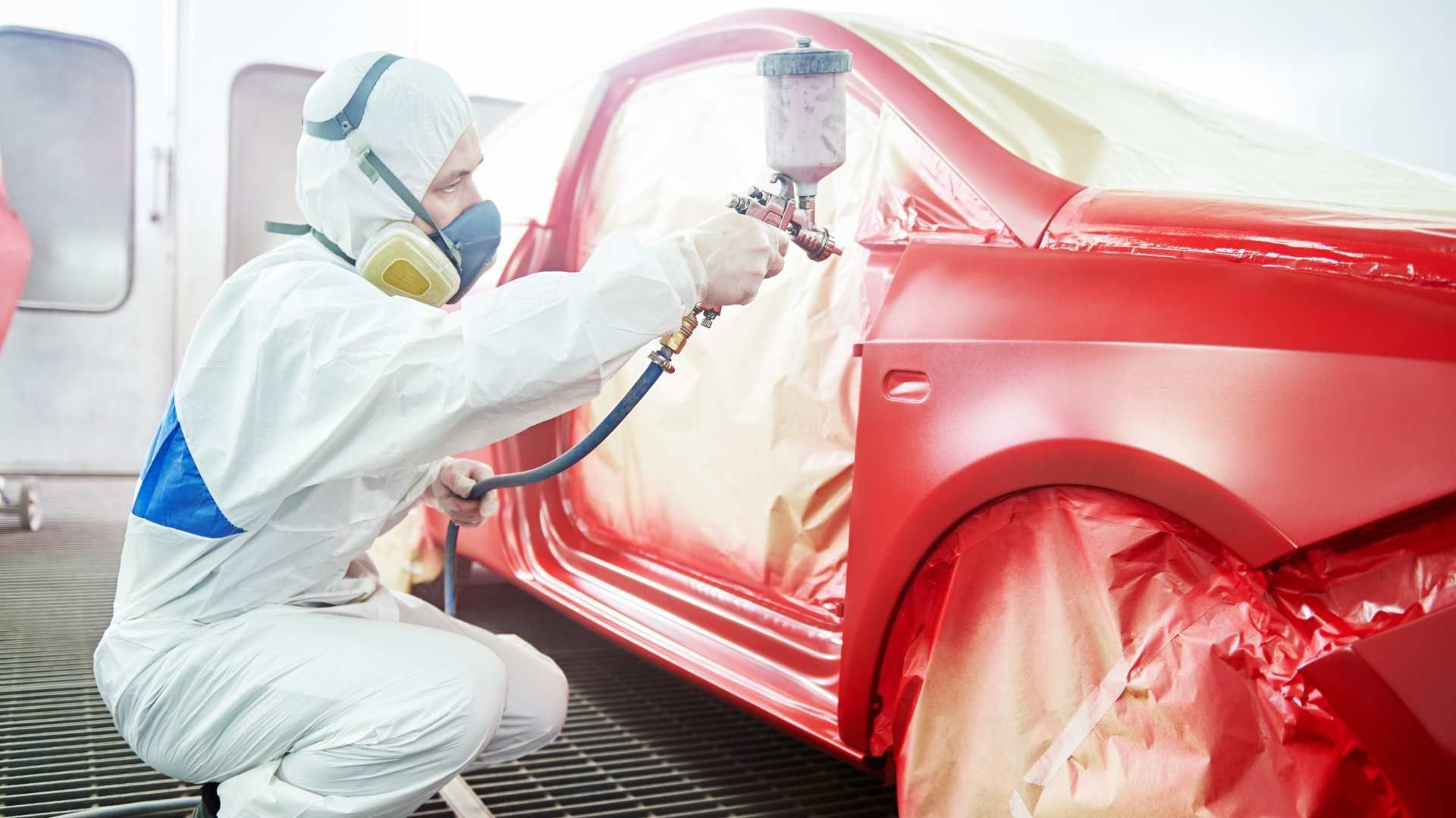 Worker spray-painting a red car in a body shop, wearing a protective suit and mask.