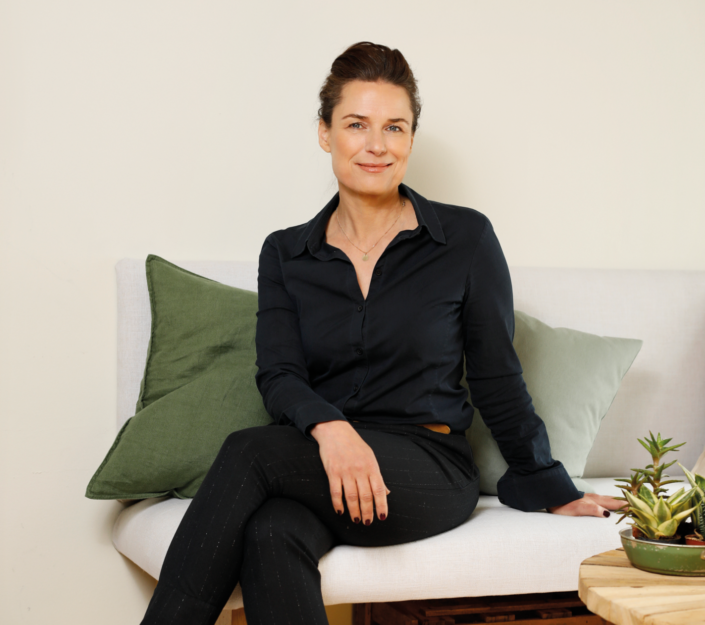Frau in schwarzem Hemd und Hose, die lächelnd auf einem Sofa mit grünen Kissen sitzt.