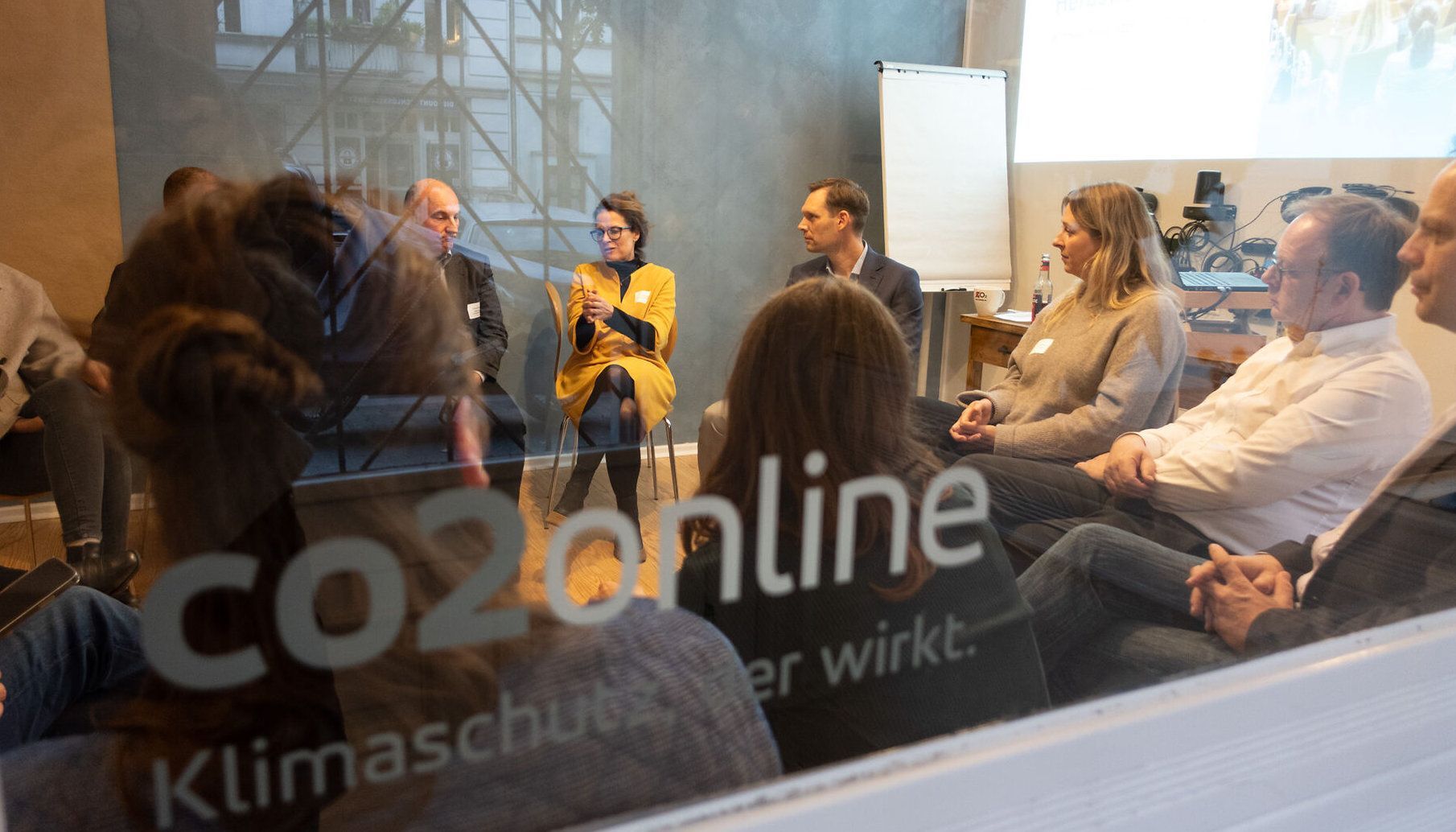 Zwei Frauen arbeiten zusammen und kleben Haftnotizen an ein Fenster; Pflanzen rahmen den Vordergrund ein.