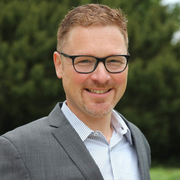 Ein lächelnder Mann mit Brille und grauem Blazer steht im Freien vor einem verschwommenen grünen Hintergrund.