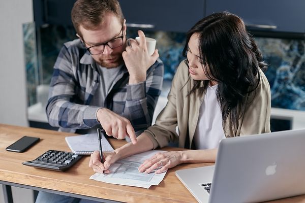 Ein Paar sitzt an einem Tisch mit Laptop, Taschenrechner und Smartphone und geht Dokumente durch. Der Mann zeigt auf etwas, während die Frau schreibt.