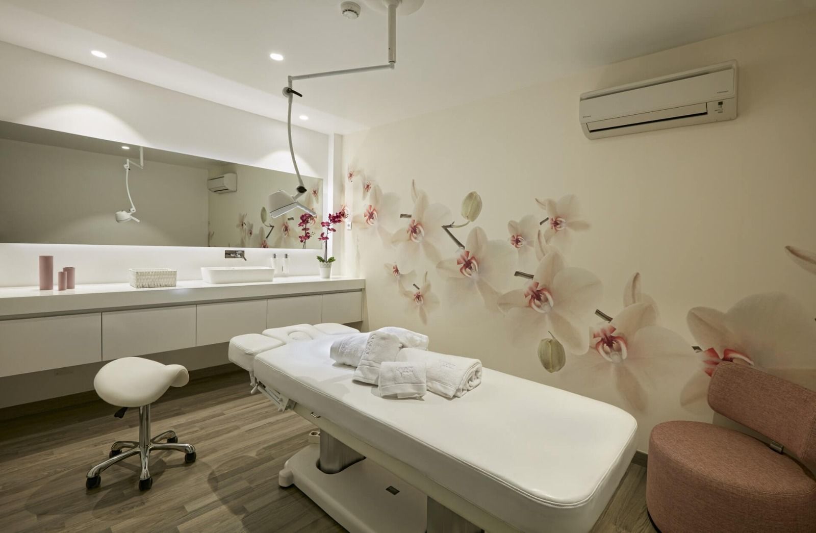 Una sala de tratamiento de spa con una pared con temática de orquídeas, mesa de tratamiento, lavabo y una silla.