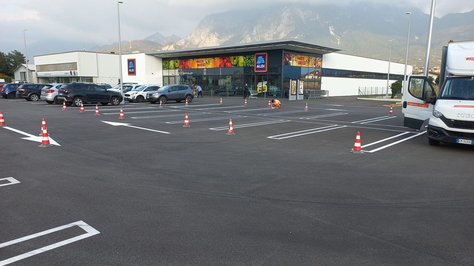 Negozio ALDI con auto nel parcheggio, coni arancioni e montagne sullo sfondo.