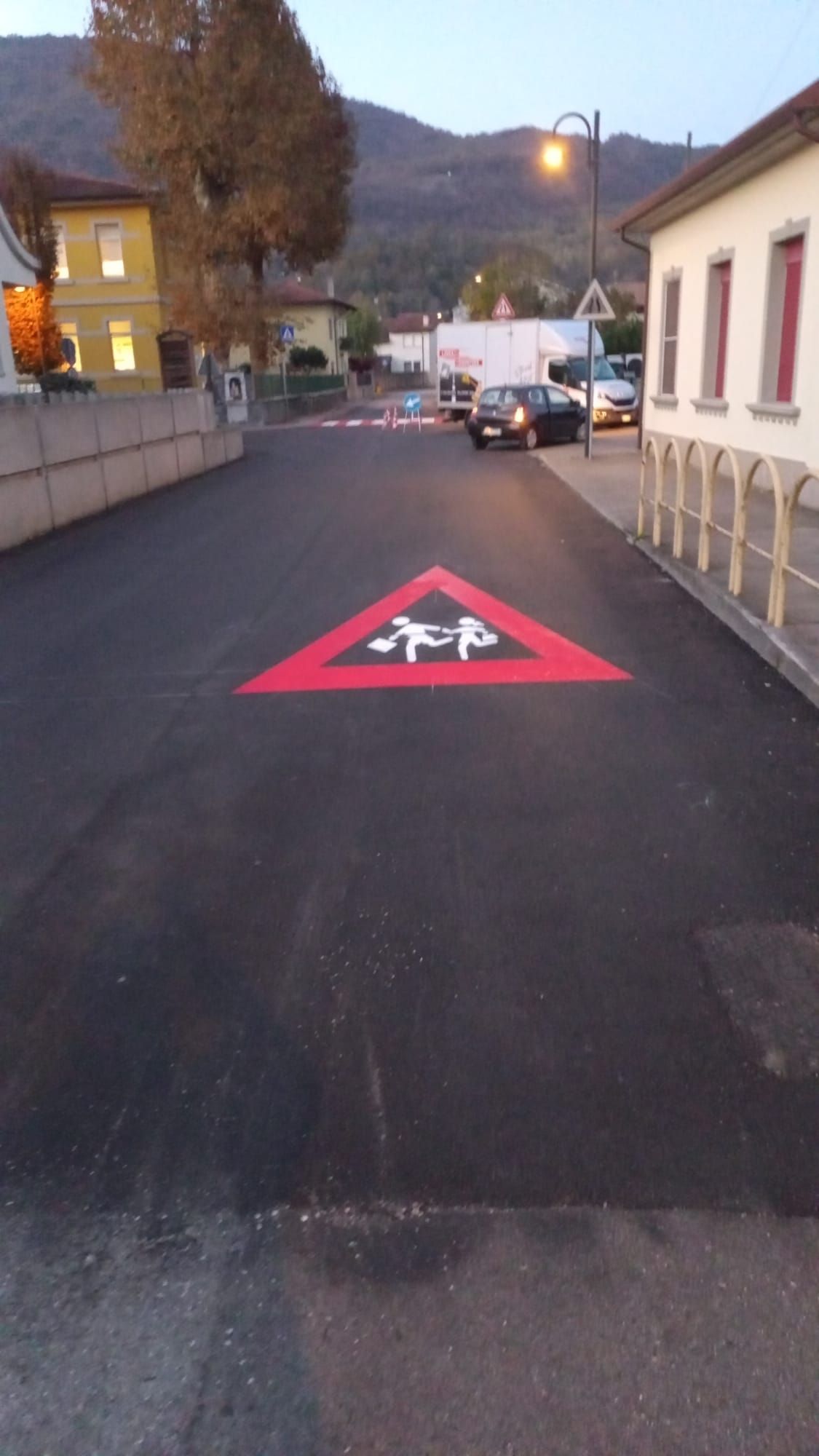 Strada con cartello di avvertimento a forma di triangolo rosso che mostra bambini che giocano. Sono visibili edifici, auto e montagne.