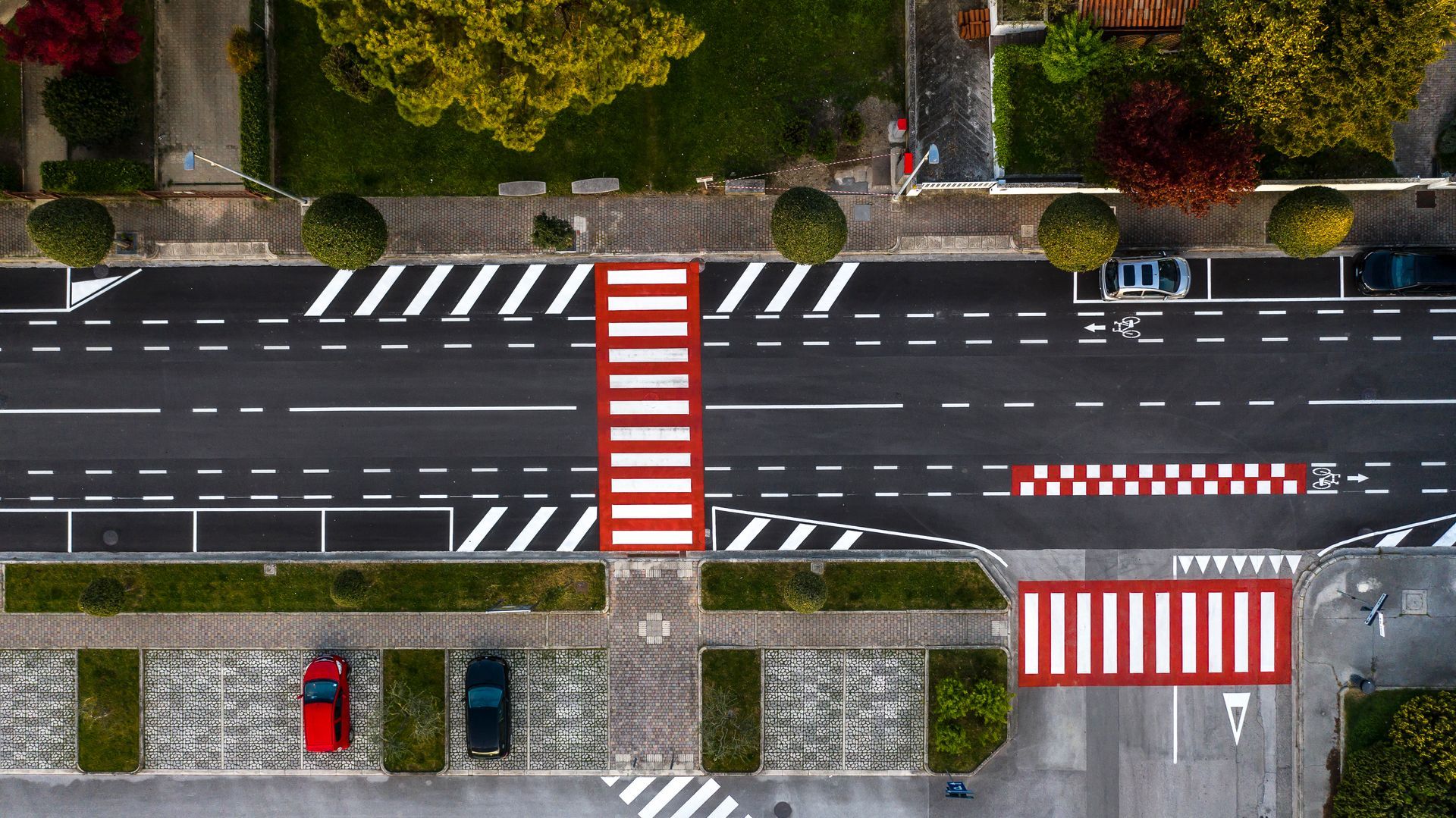 Vista dall'alto di una strada con attraversamenti pedonali, parcheggi e spazi verdi.