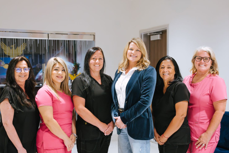 Six women standing together indoors, smiling in front of a blue abstract wall display.