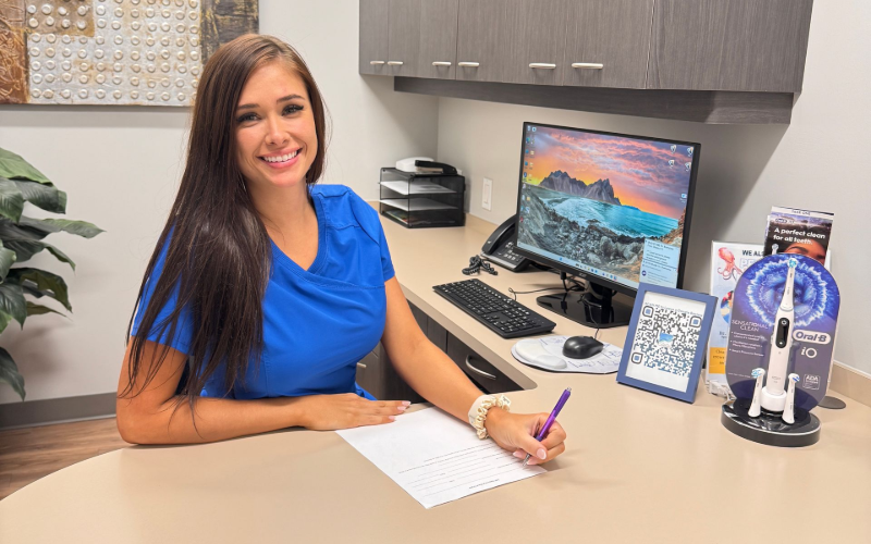 A person in a blue scrub top sitting at a desk and writing on paper in an office setting.