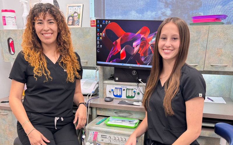 Two professionals in black scrubs smiling in an office setting with a computer monitor in the background.