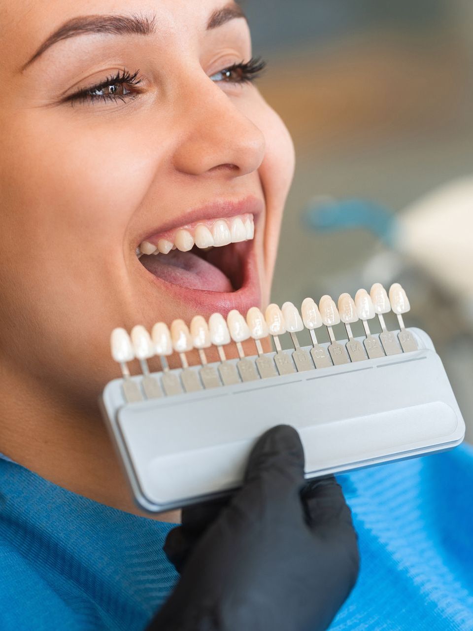 A woman is smiling while holding a tooth color chart in front of her mouth.