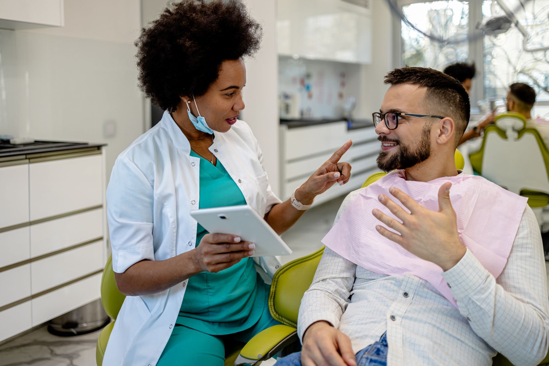 A dentist is talking to a patient in a dental chair while holding a tablet.
