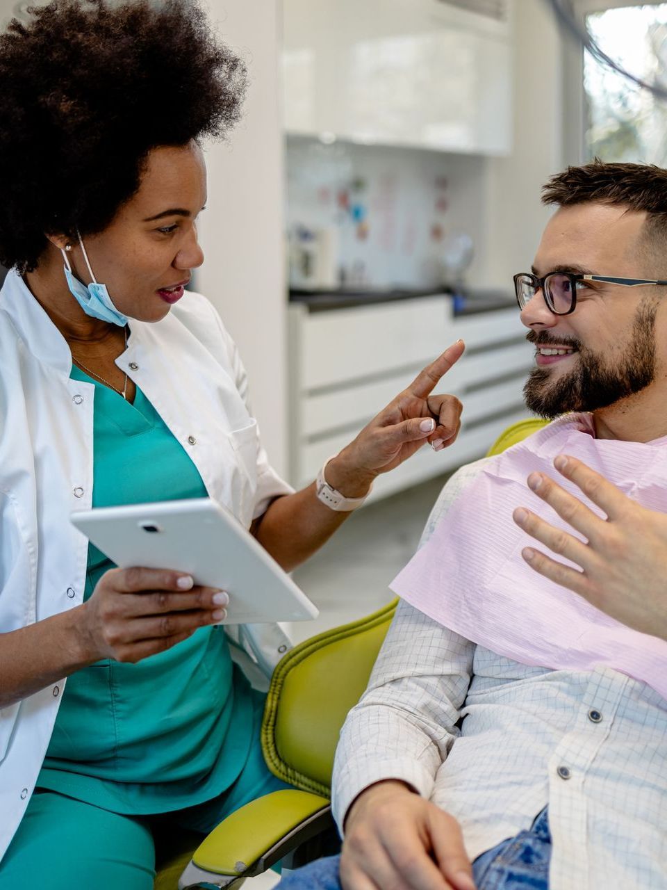 A dentist is talking to a patient in a dental chair while holding a tablet.