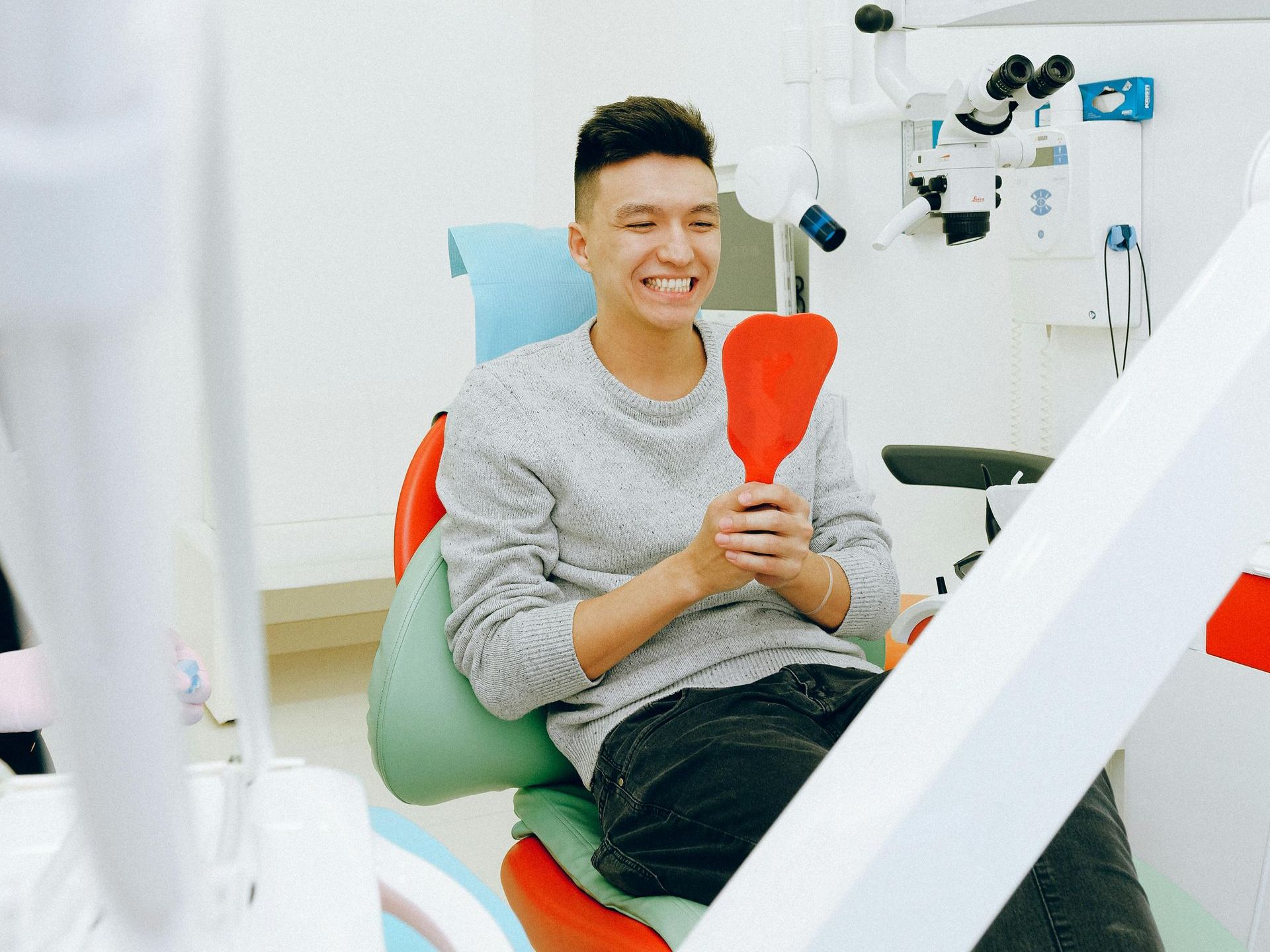 A man is sitting in a dental chair looking at his teeth in a mirror.