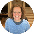 Woman with curly brown hair smiles, wearing a blue sweater. Interior setting with stairs visible.