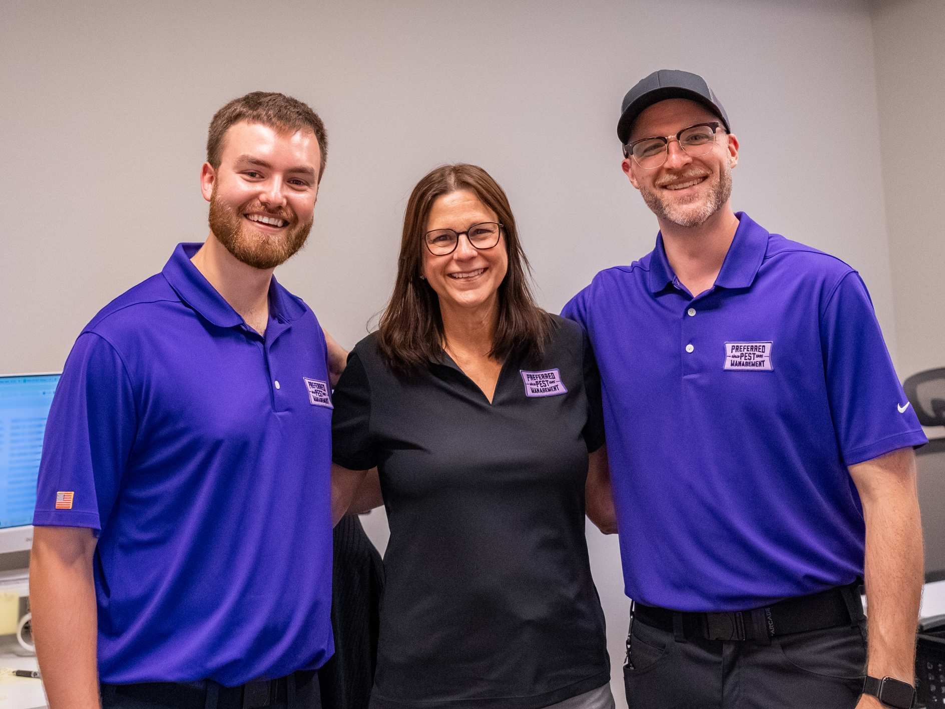 Three smiling coworkers in purple and black polos posing indoors against a plain wall