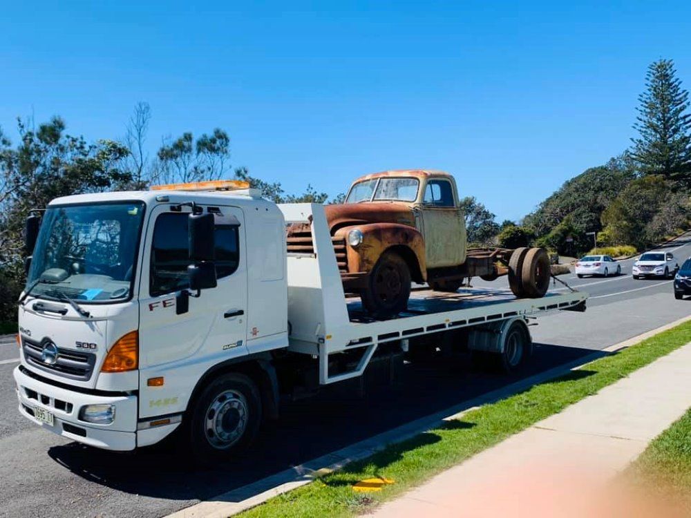 A White Tow Truck is Carrying an Old Truck on the Back of It — Port Macquarie Tilt Tray Towing in Wauchope, NSW