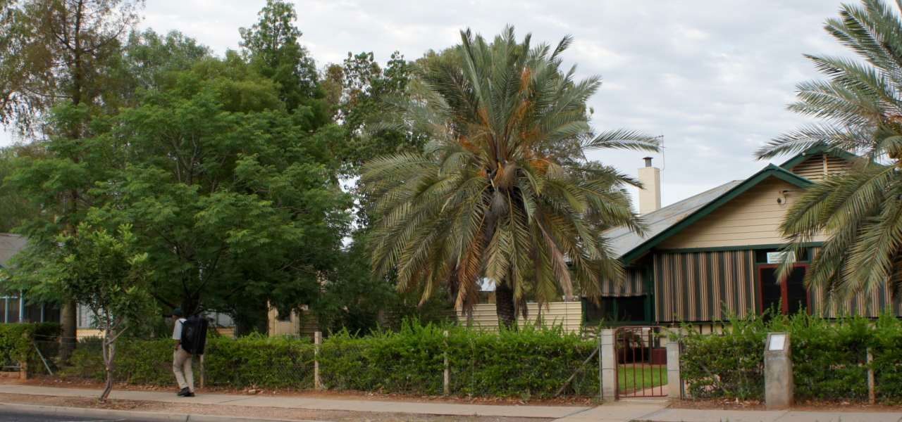 A man is walking down a sidewalk in front of a house with palm trees in the background.