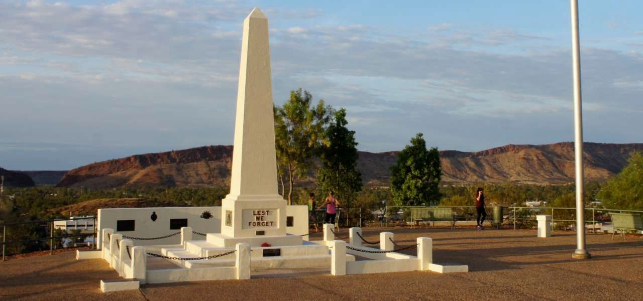 A white obelisk is in the middle of a park with mountains in the background.