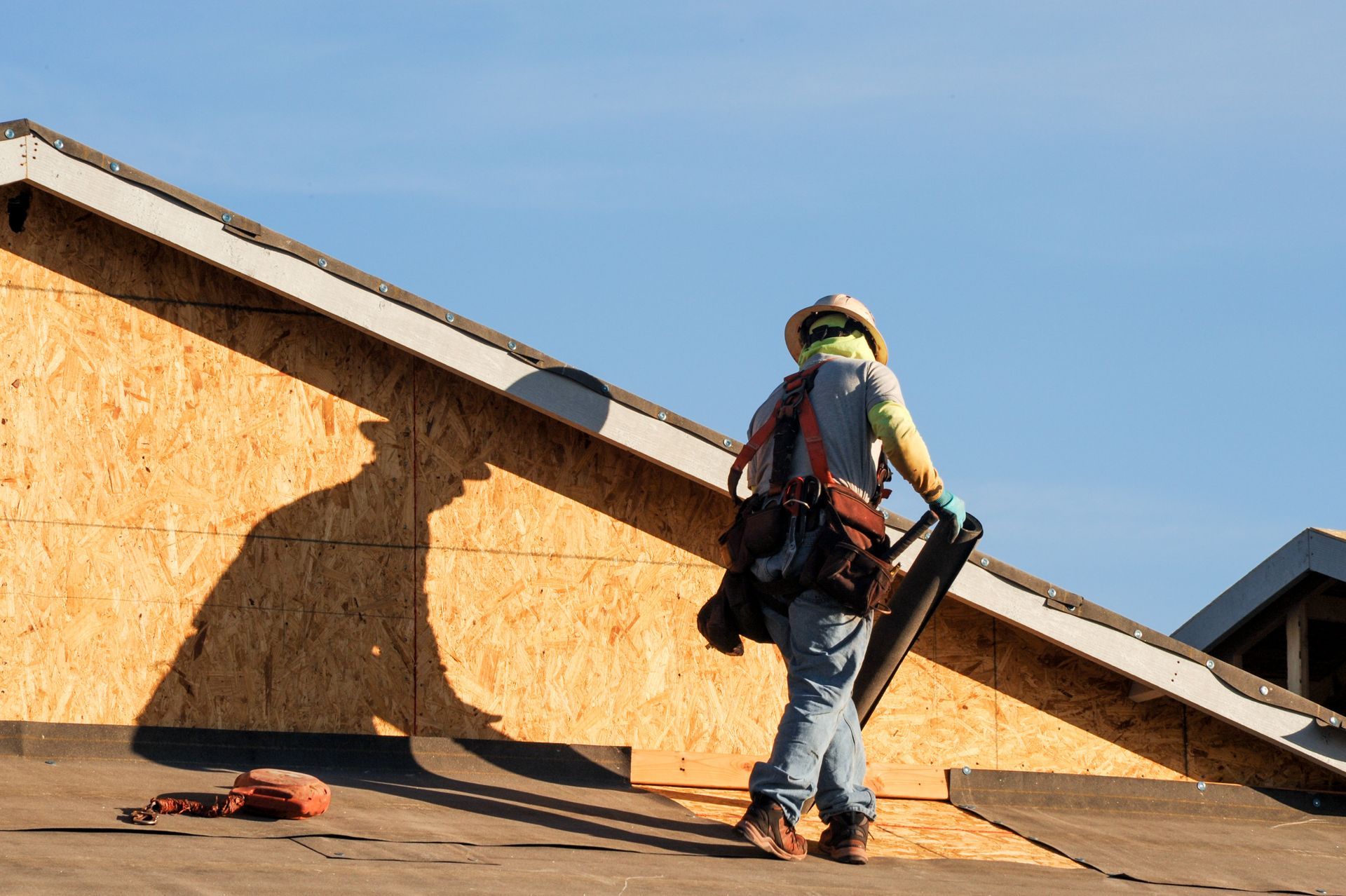 Worker installing roofing on a plywood-covered roof under a clear blue sky