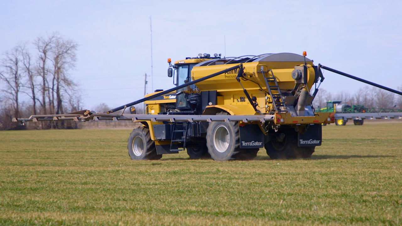 A bright yellow agricultural sprayer moves across a large, flat green field on a clear, sunny day.