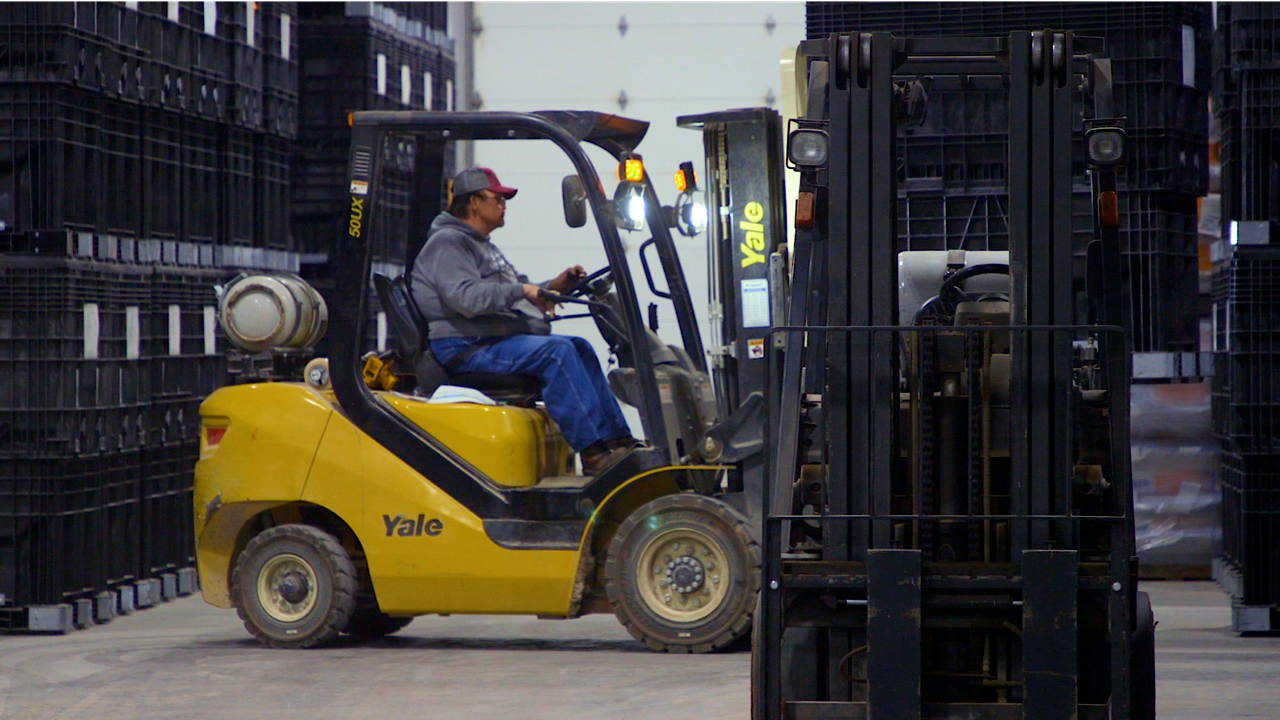 A person wearing a grey hoodie drives a yellow Yale forklift in a warehouse aisle between stacks of black crates.