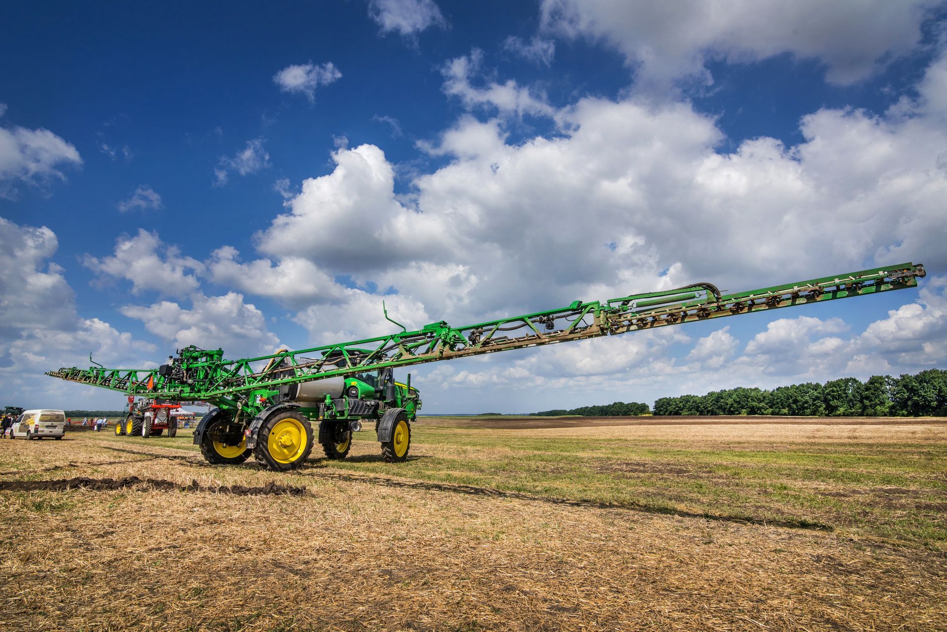 A green agricultural sprayer with long, extended booms sits in a dry, harvested field under a bright, cloudy sky.