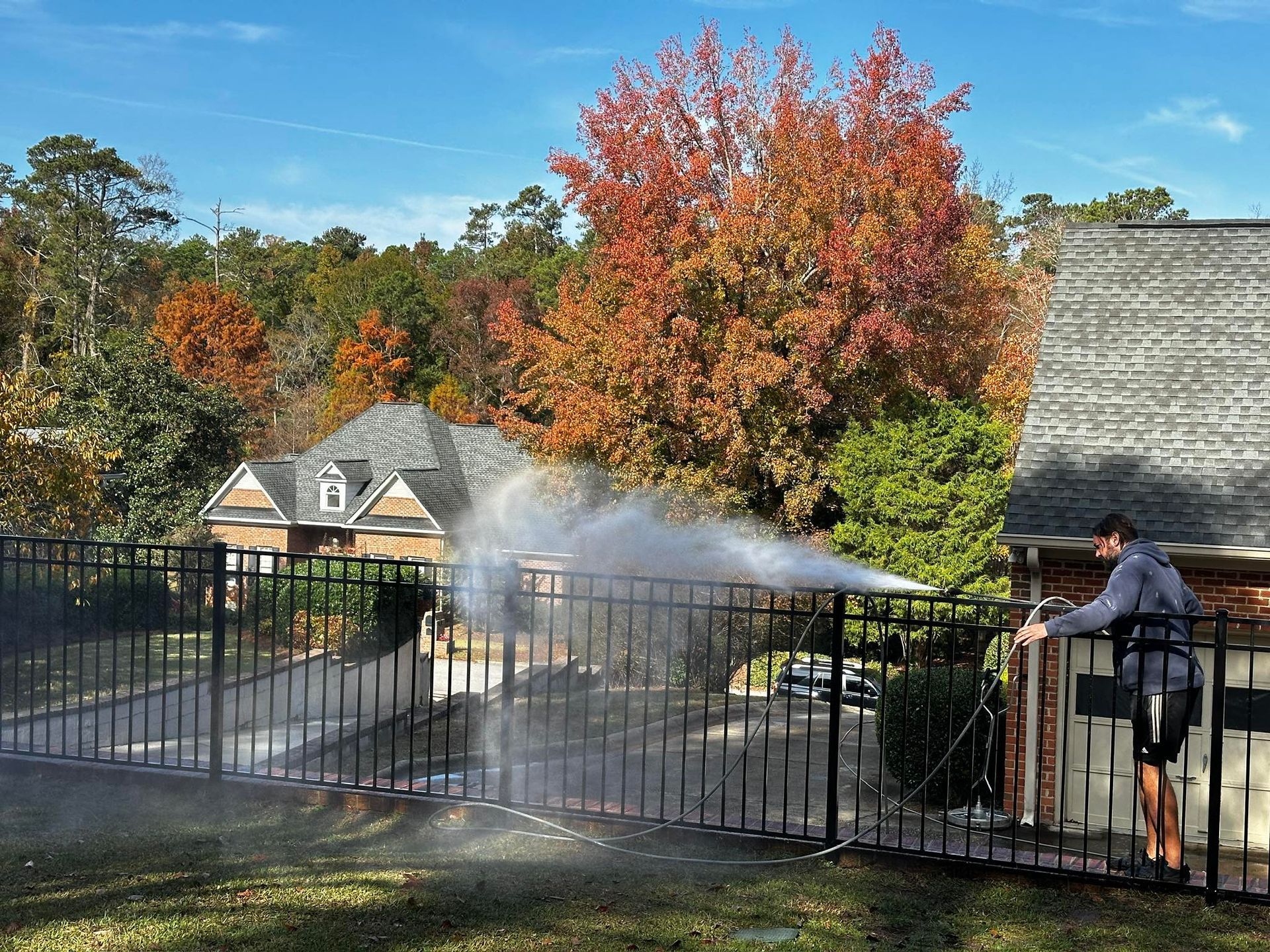 Person sprays water over a fenced yard with autumn trees and houses in the background.
