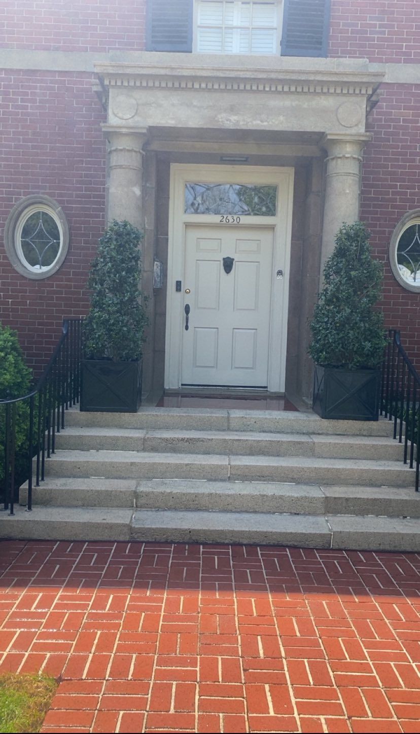 Brick building entrance with white door, stone steps, and oval windows, framed by tall shrubs.