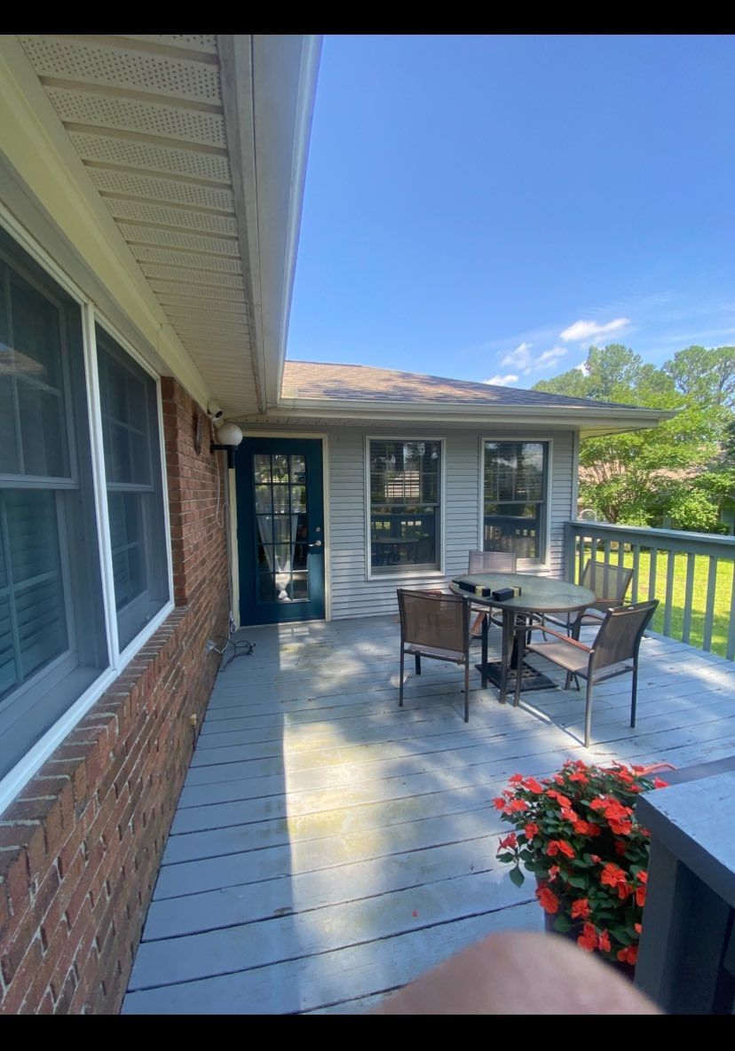 Covered backyard patio with dining table, chairs, and potted flowers beside a brick house under a blue sky