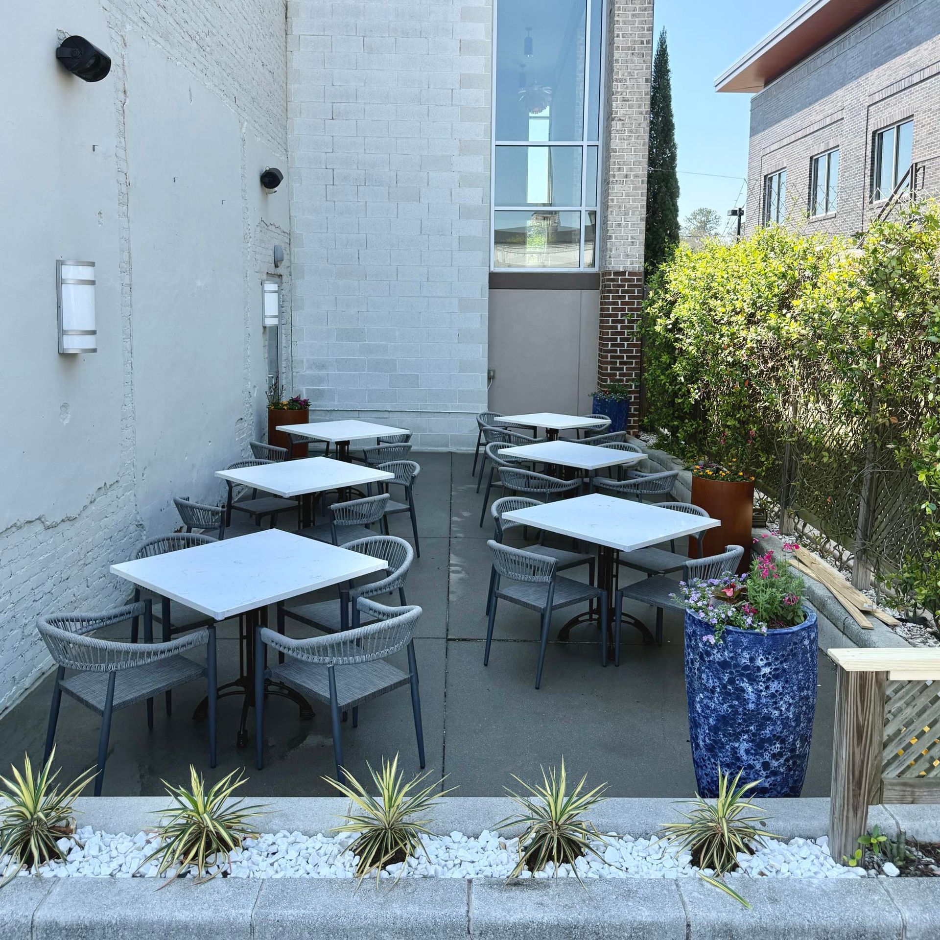 Small outdoor cafe patio with white tables and gray chairs beside a white building and greenery.
