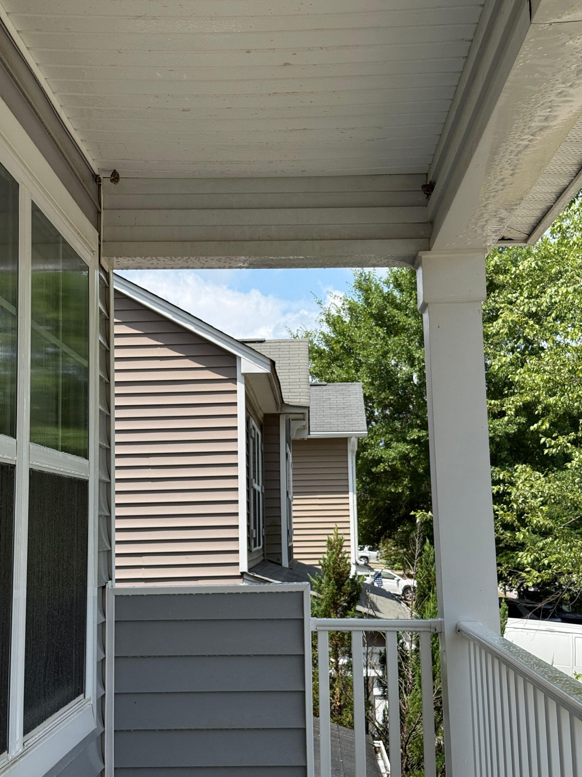 Covered apartment balcony with white railing, tan siding, and trees beyond the porch.