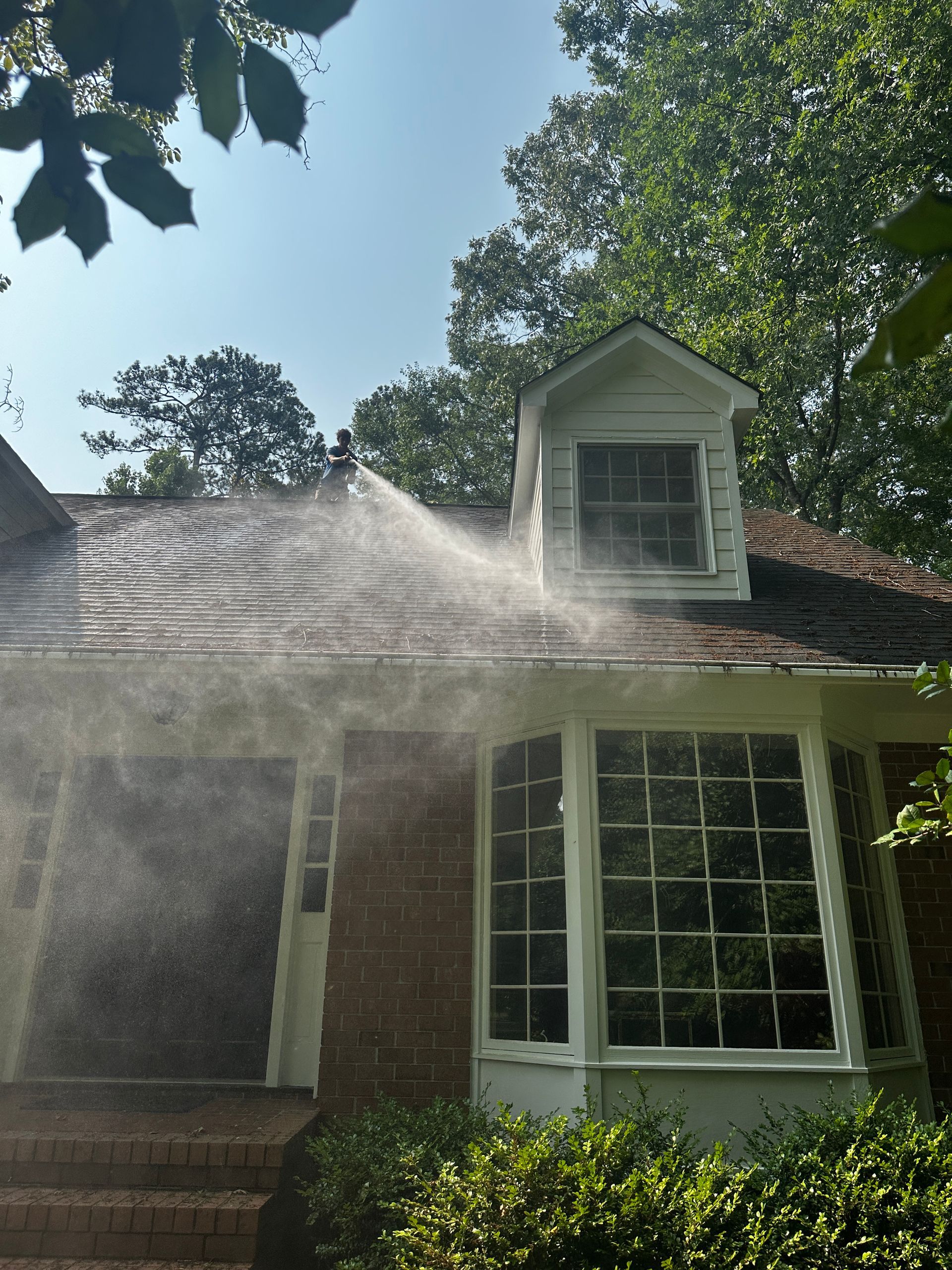 Water spraying across a red brick house roof with dormer windows and a sunlit front yard
