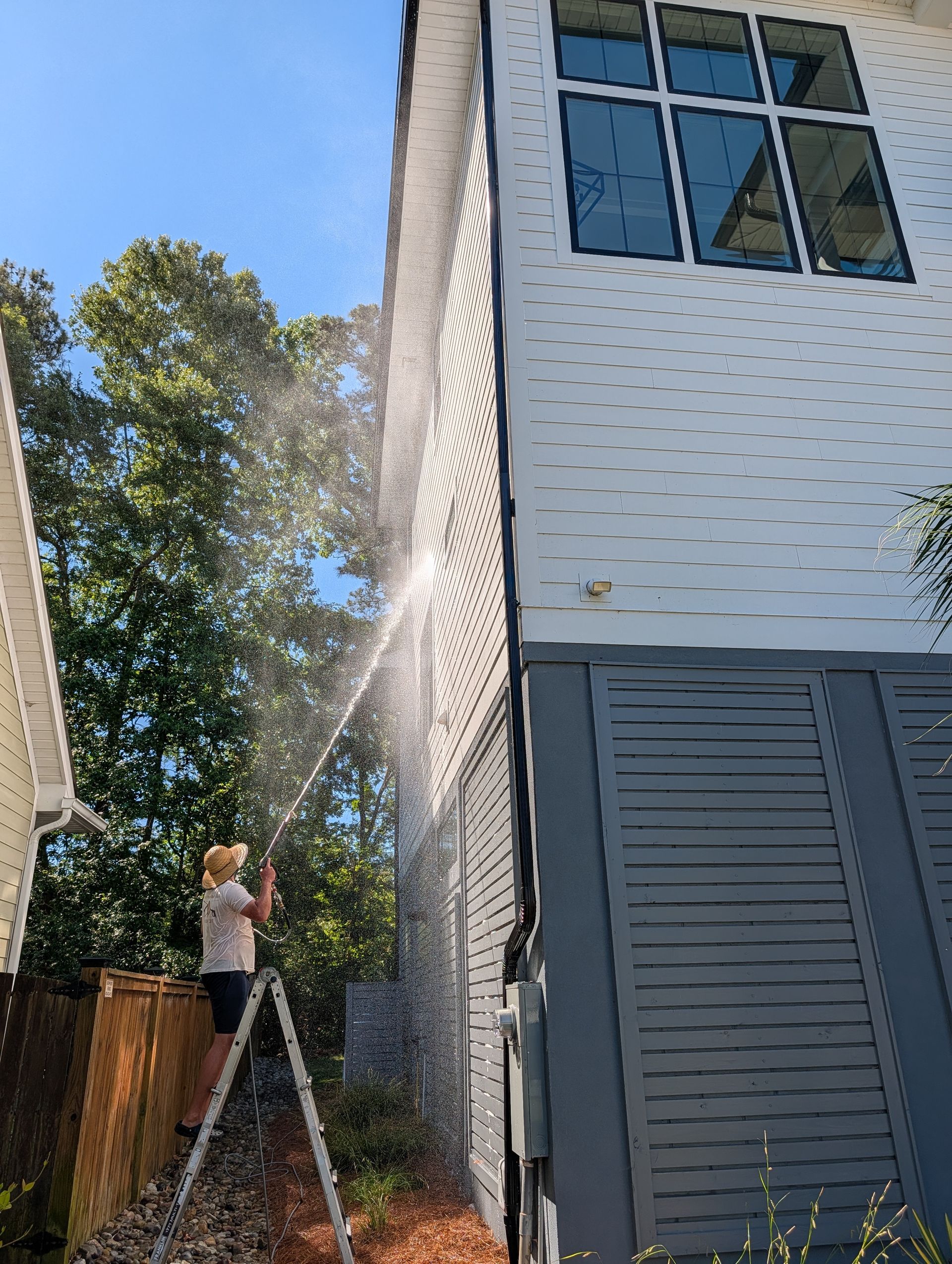 Person on a ladder pressure-washing the side of a two-story house beside trees and a fence