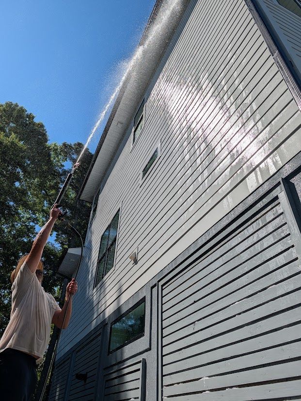 Person on a ladder cleaning a white house exterior with a long brush under a blue sky