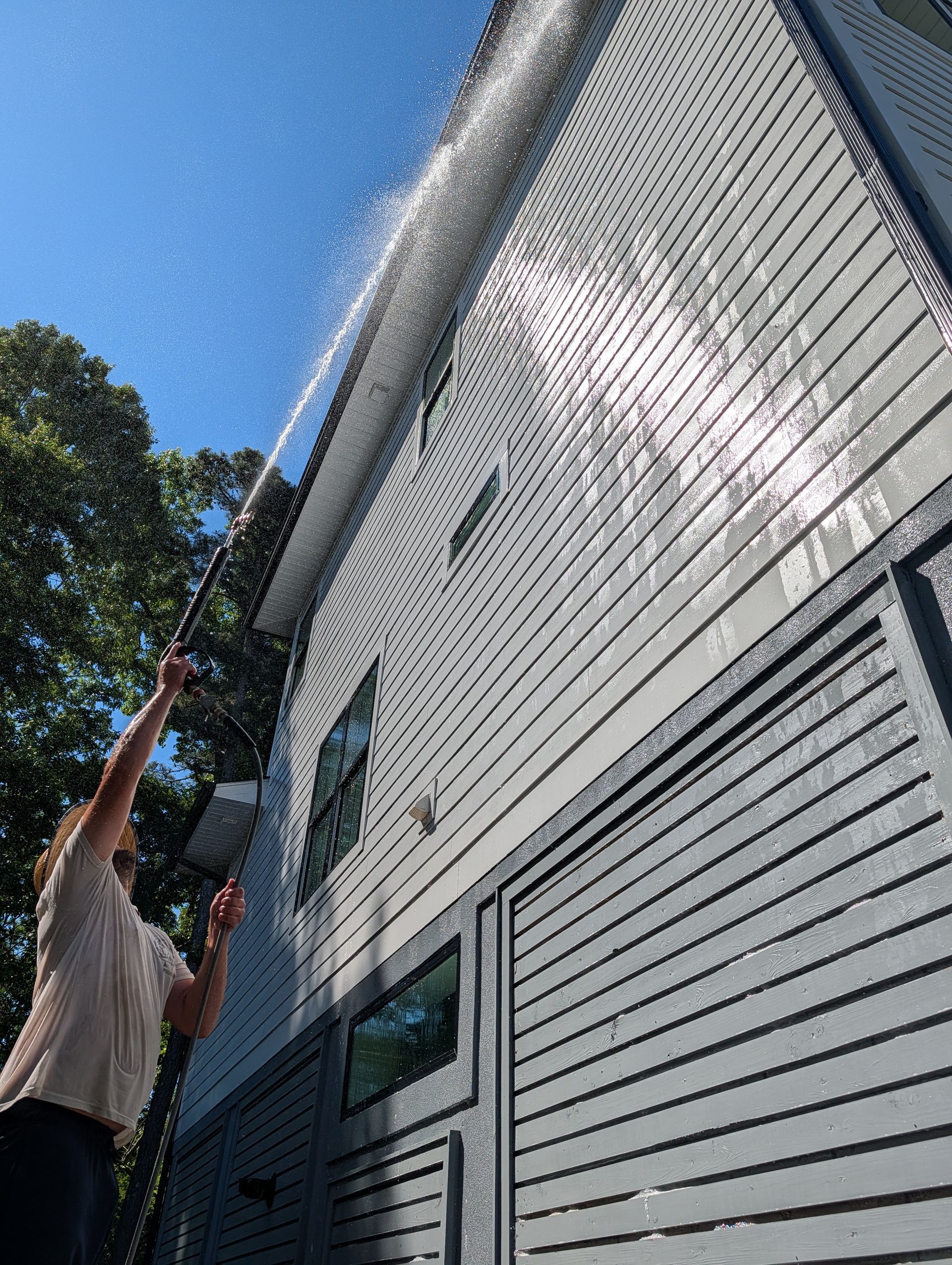 Person using a long pole to clean a white-sided house under a blue sky