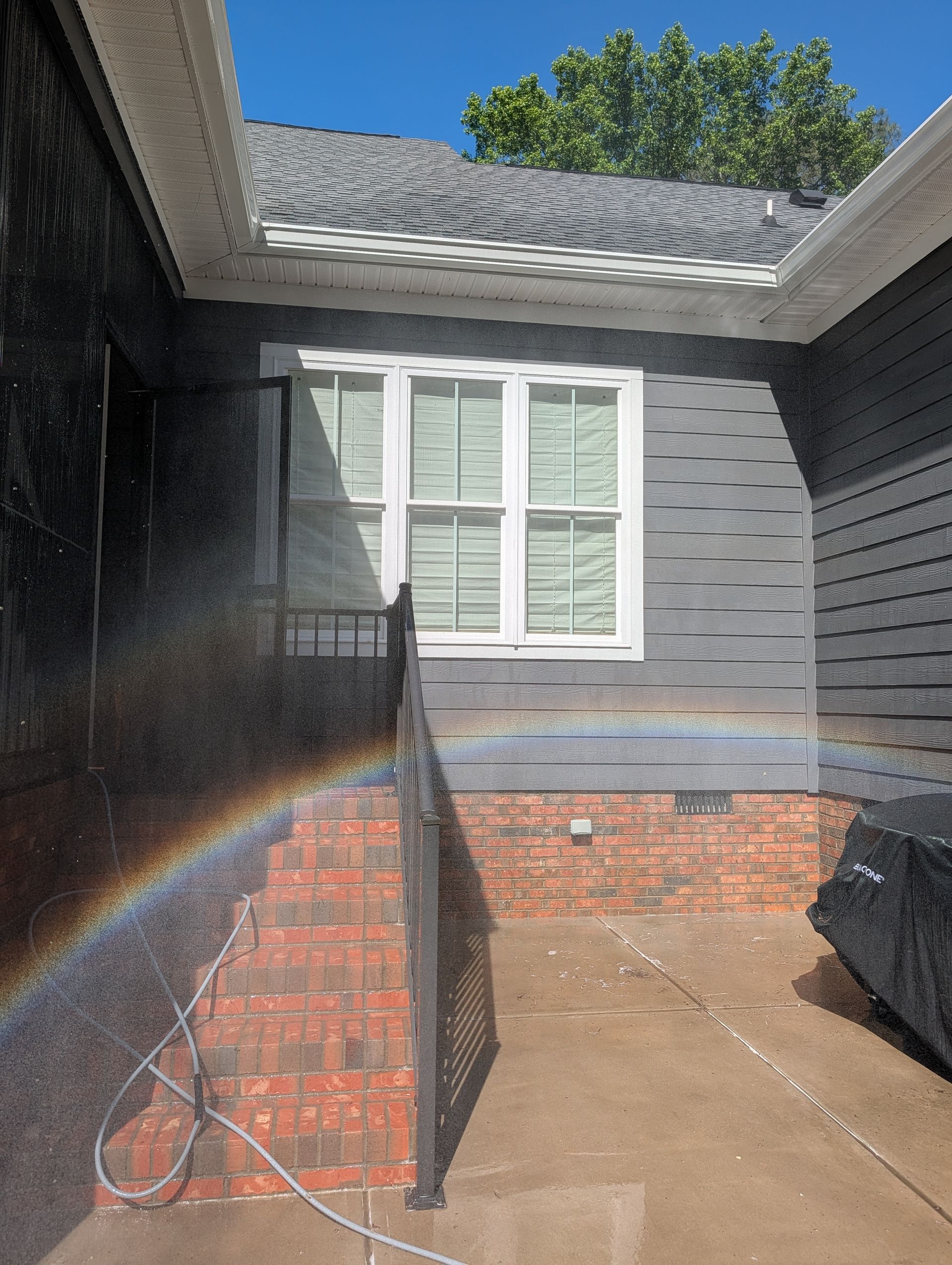 Covered front patio with brick columns, gray siding, white shutters, and a rainbow from sunlight spray.
