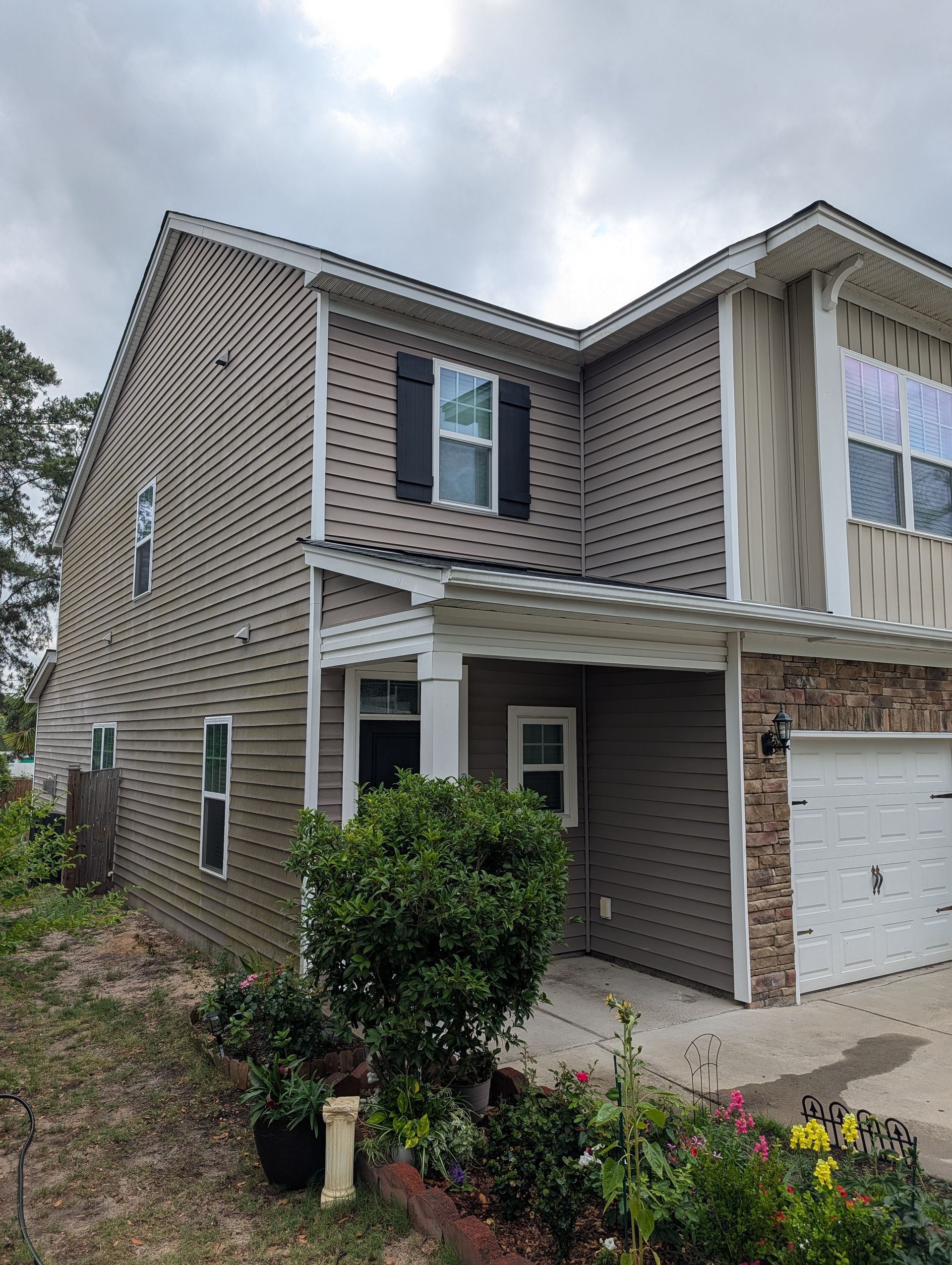 Two-story beige brick house with white trim, covered porch, garage, and flower bed under cloudy sky