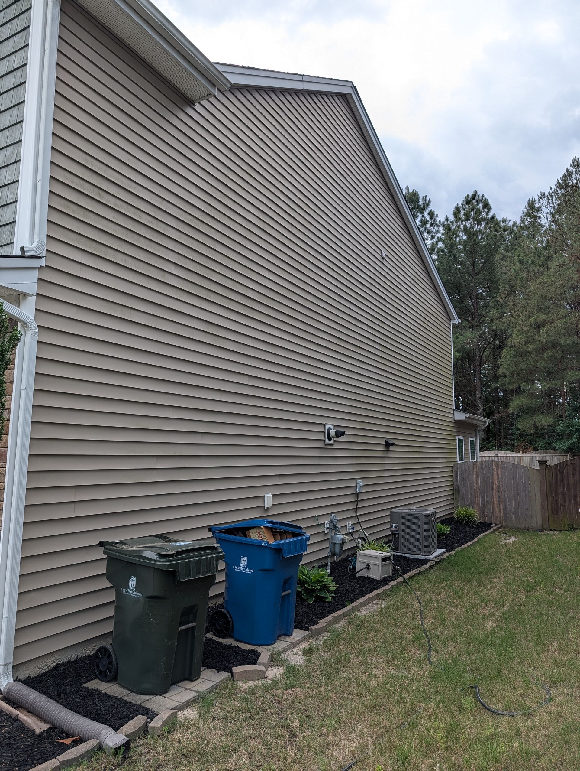 Side of beige house with trash bins, utility pipes, and a grassy yard under cloudy sky