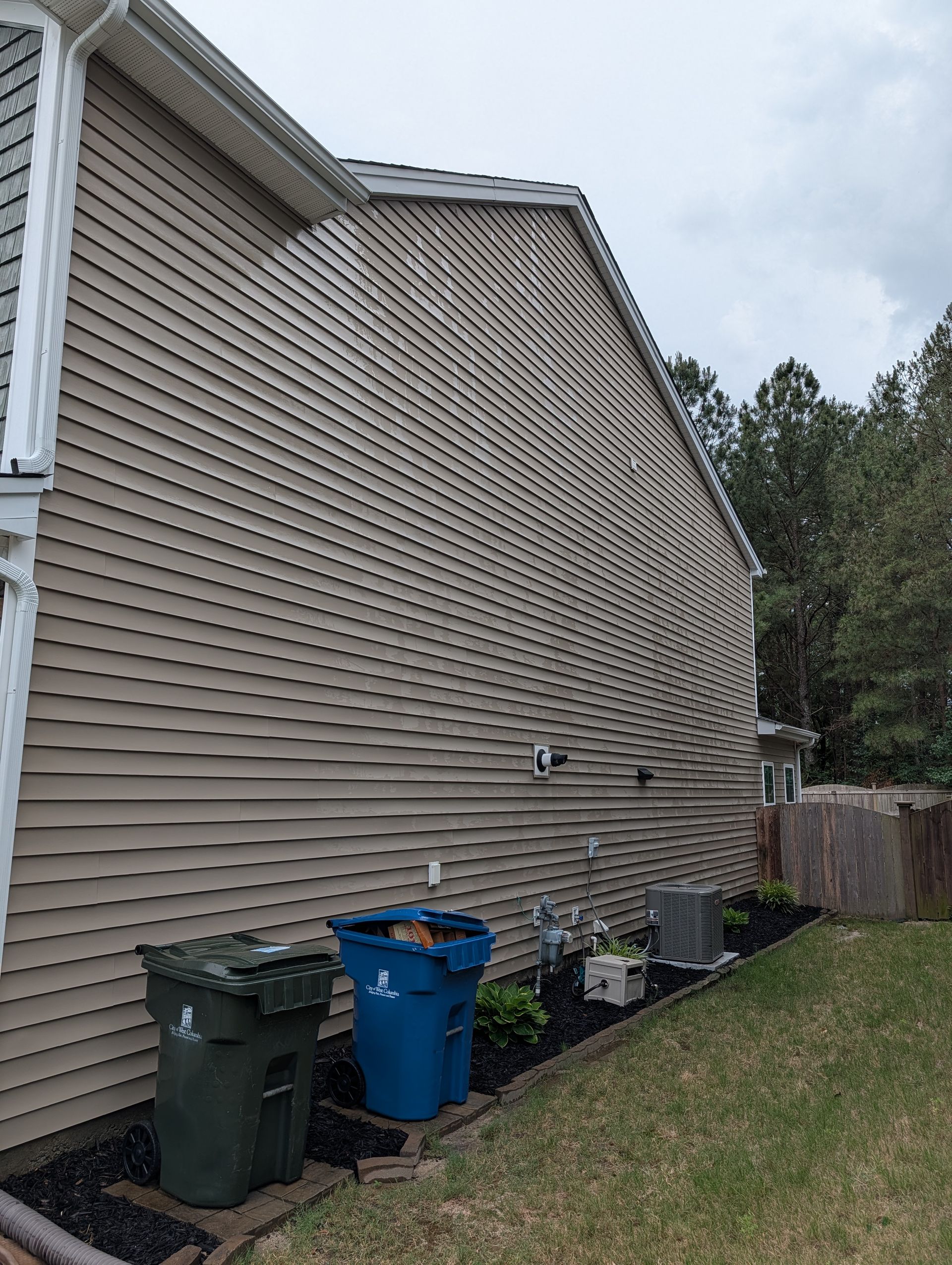 Side view of a beige house with damaged siding, trash bins, and a grassy yard under cloudy skies