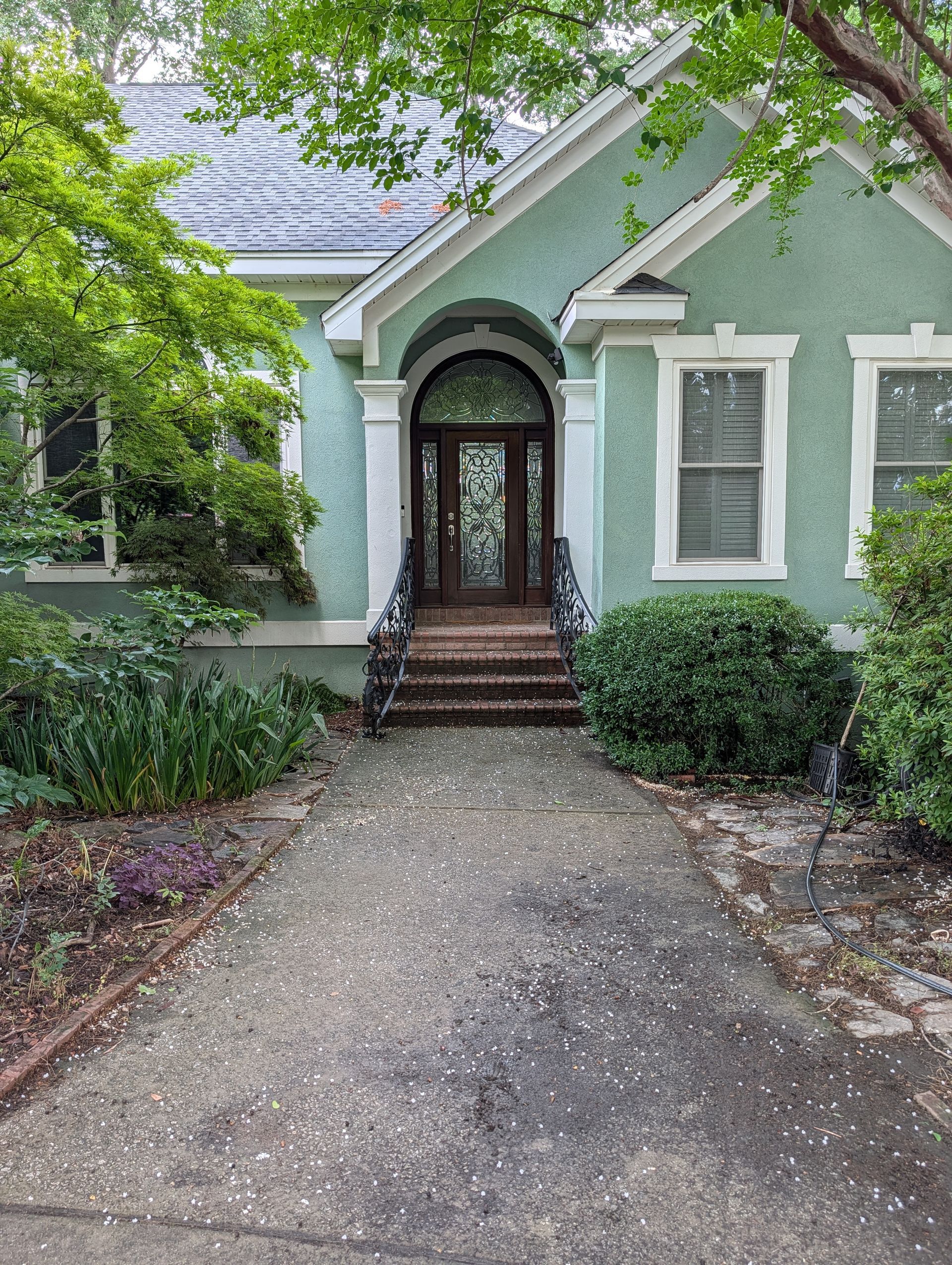 Front view of a mint-green house with an arched doorway and a gravel driveway, framed by trees and shrubs