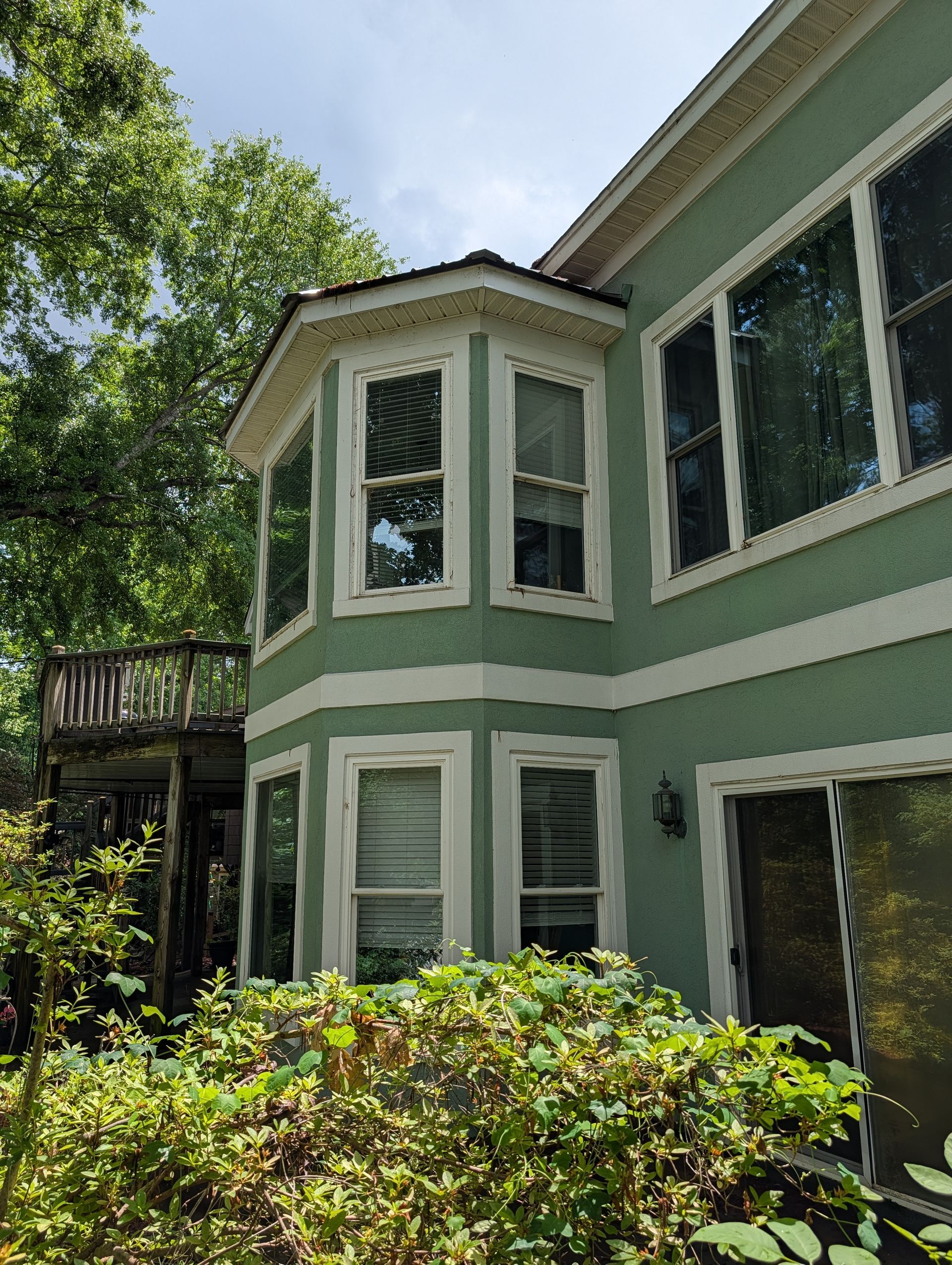 Green two-story house with bay windows, white trim, and shrubs in front under trees