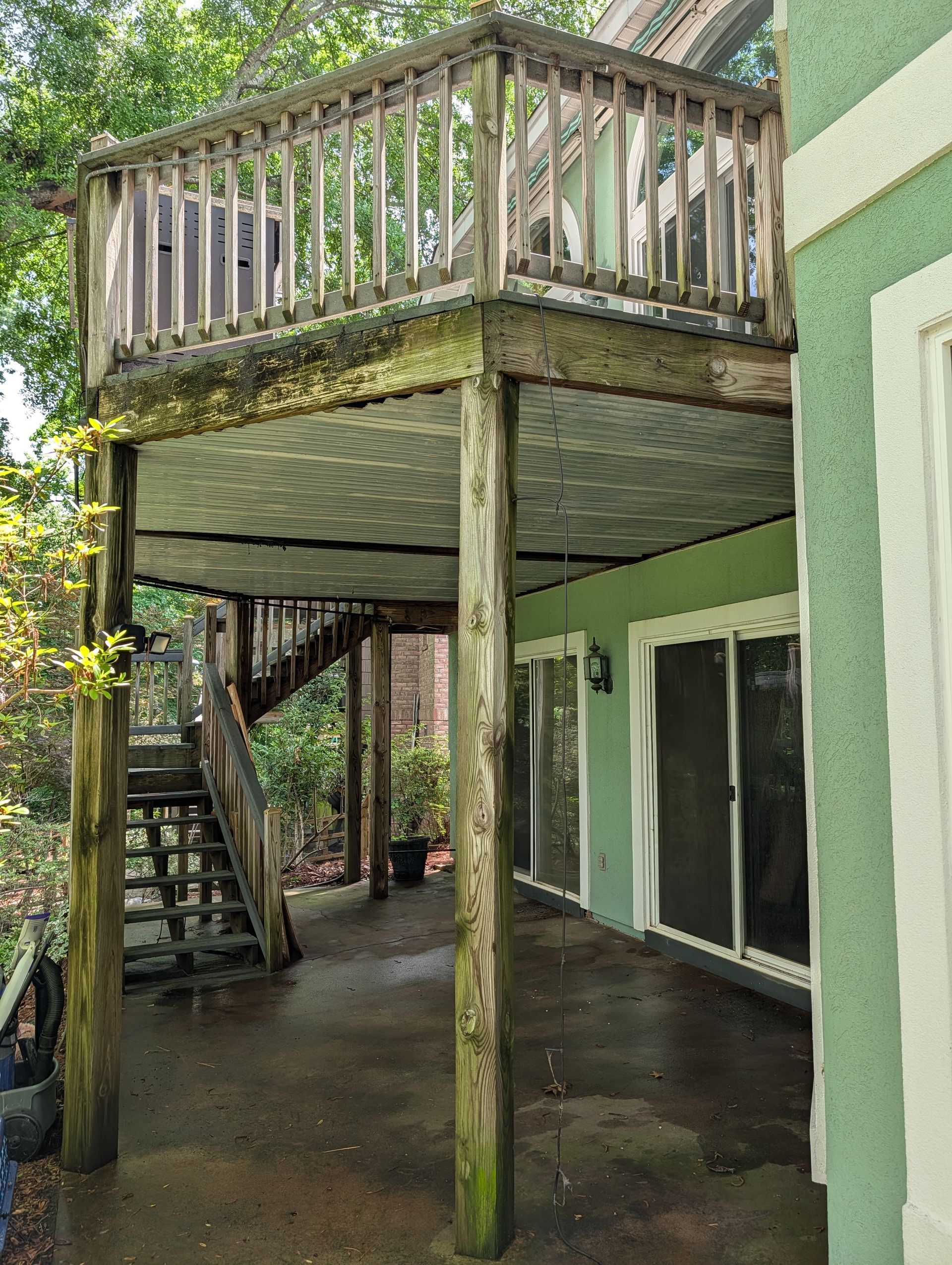 Moss-covered elevated wooden deck and staircase beside a green house, over a wet concrete patio.