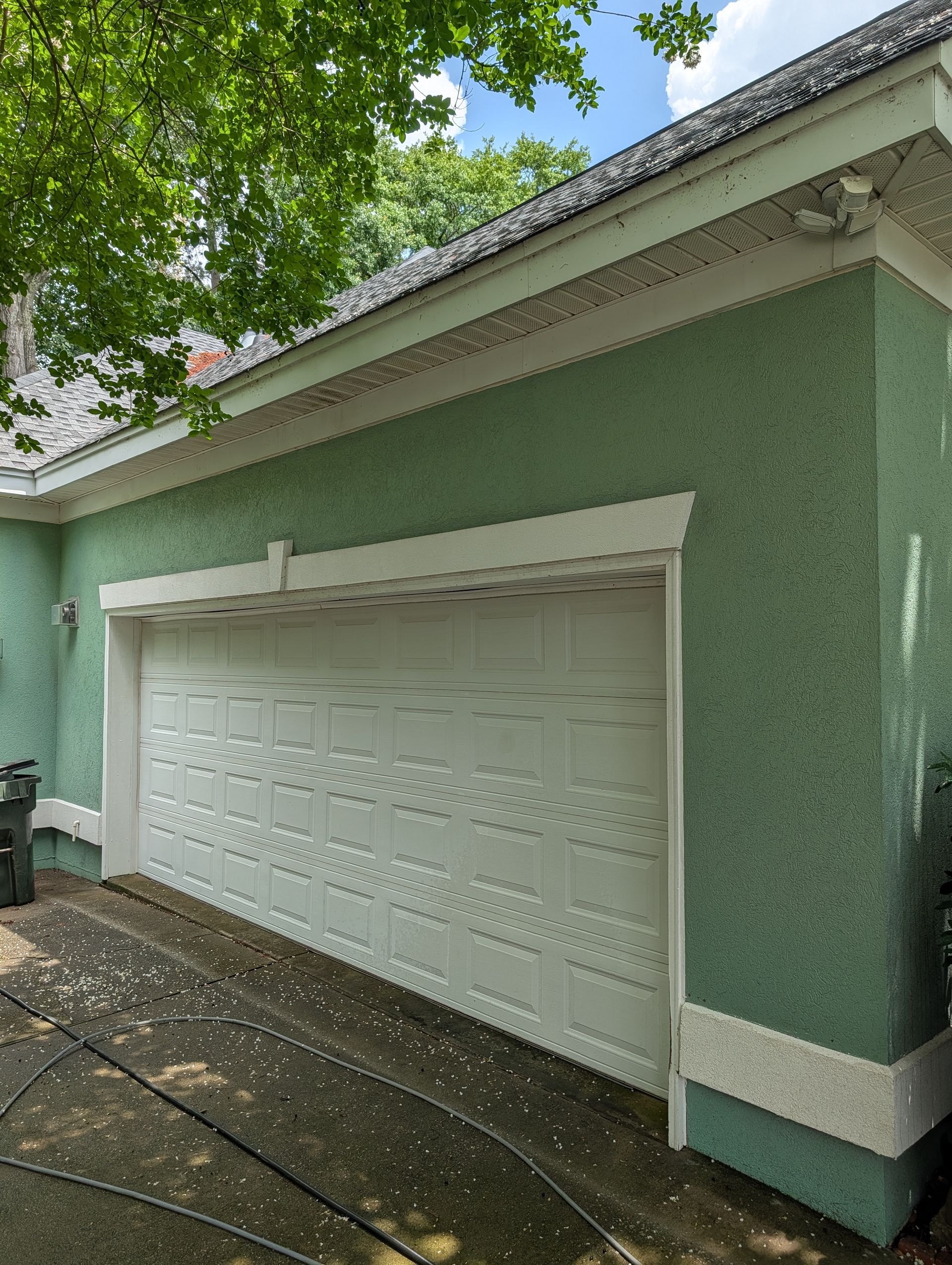 Green stucco garage with white door and trim under leafy trees beside a wet driveway
