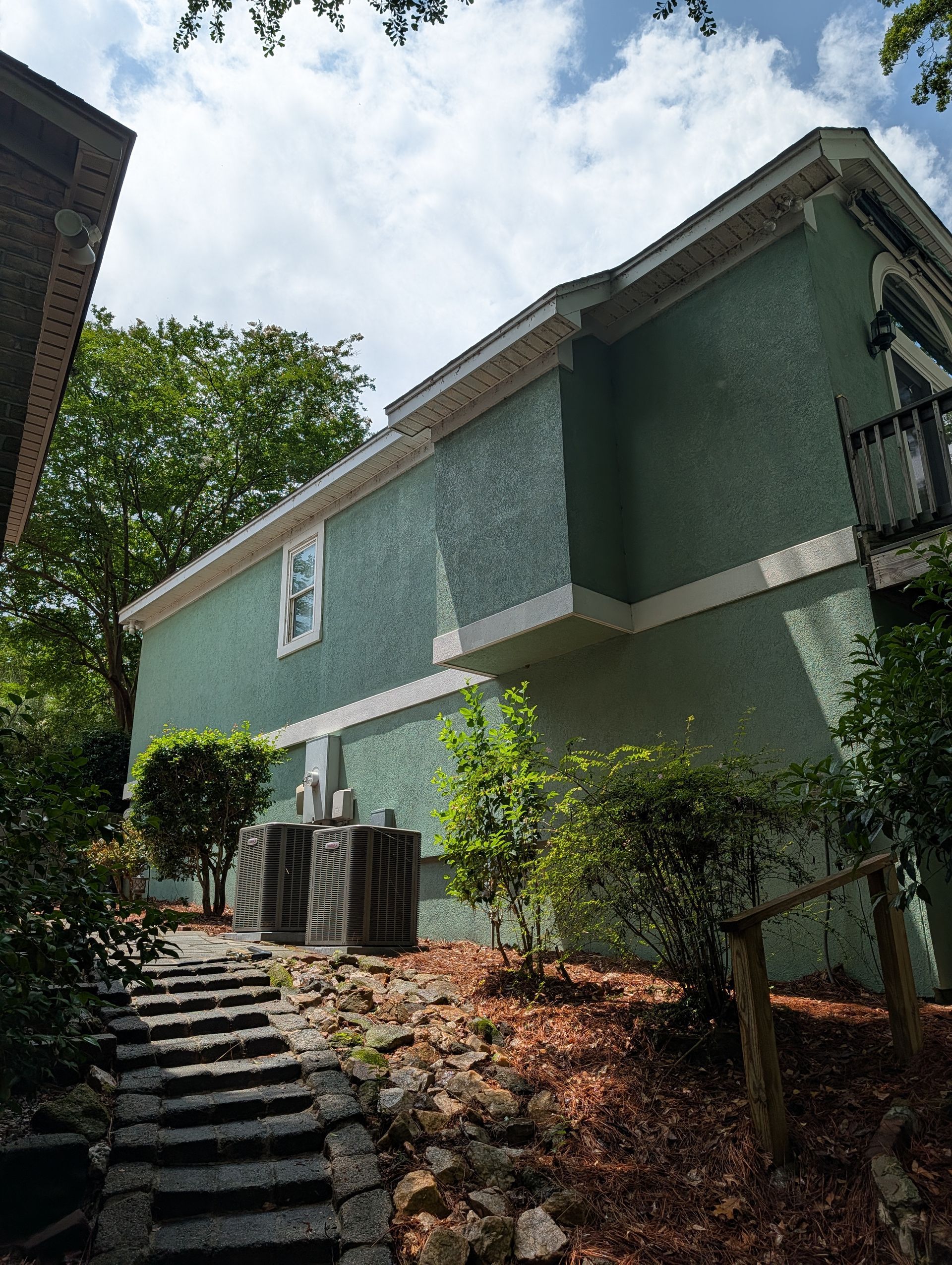 Two-story green building with stairs, trees, and outdoor AC units on a sunny hillside.