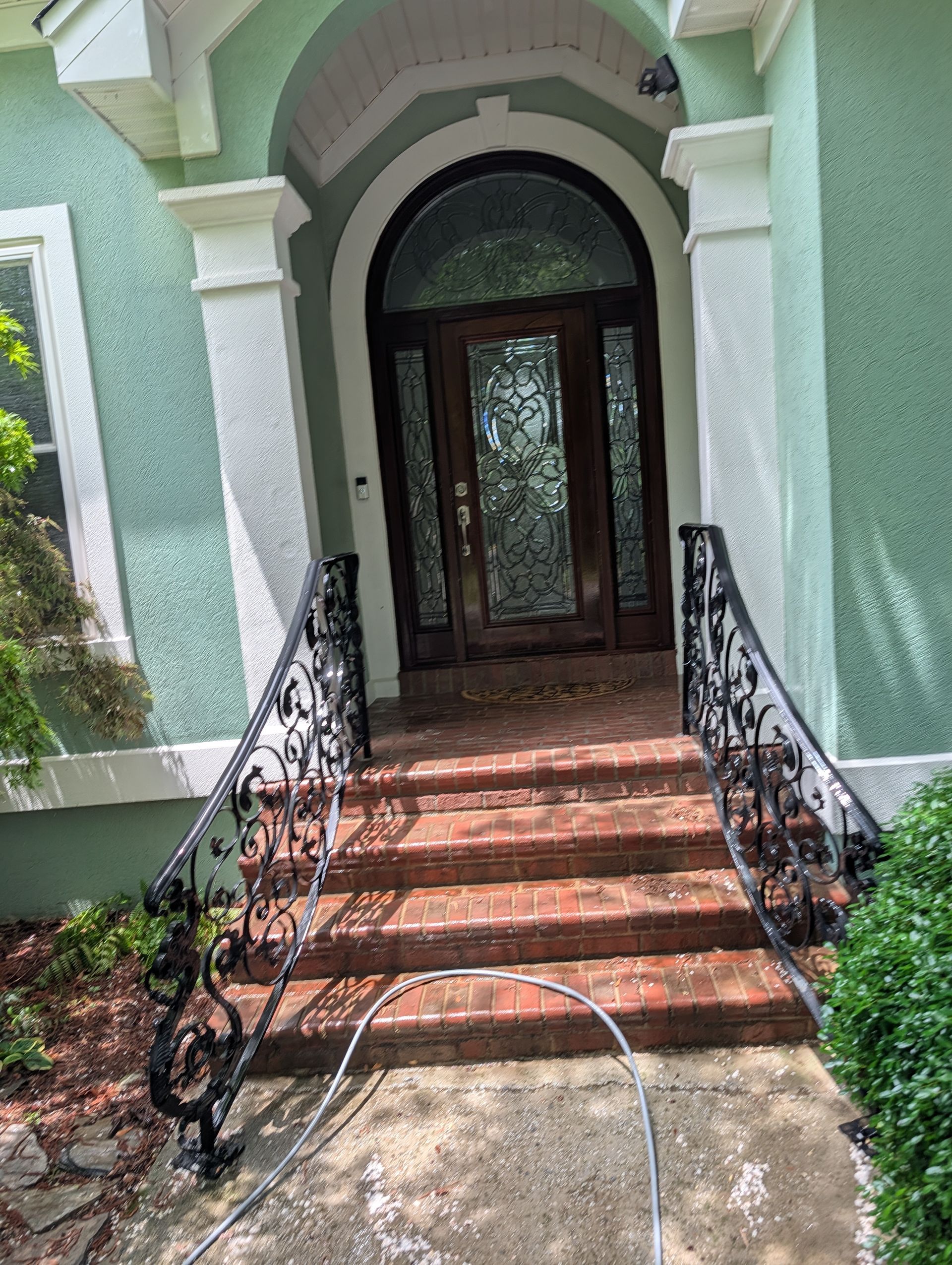 Front entrance with brick steps, wrought-iron railings, and arched wooden doors on a green house