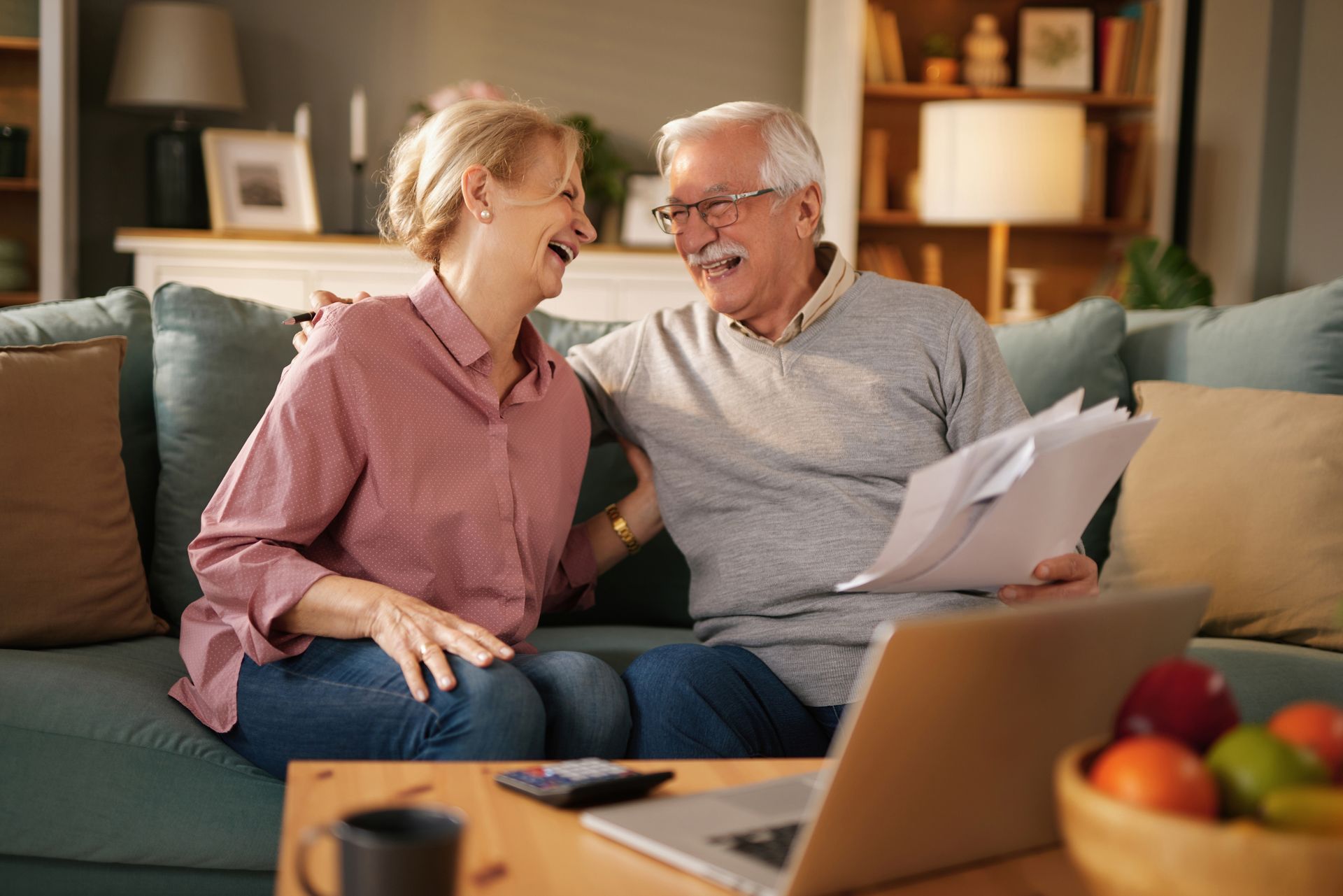 Two people smiling on a couch, reviewing papers in a cozy living room with a laptop and bowl of fruit nearby