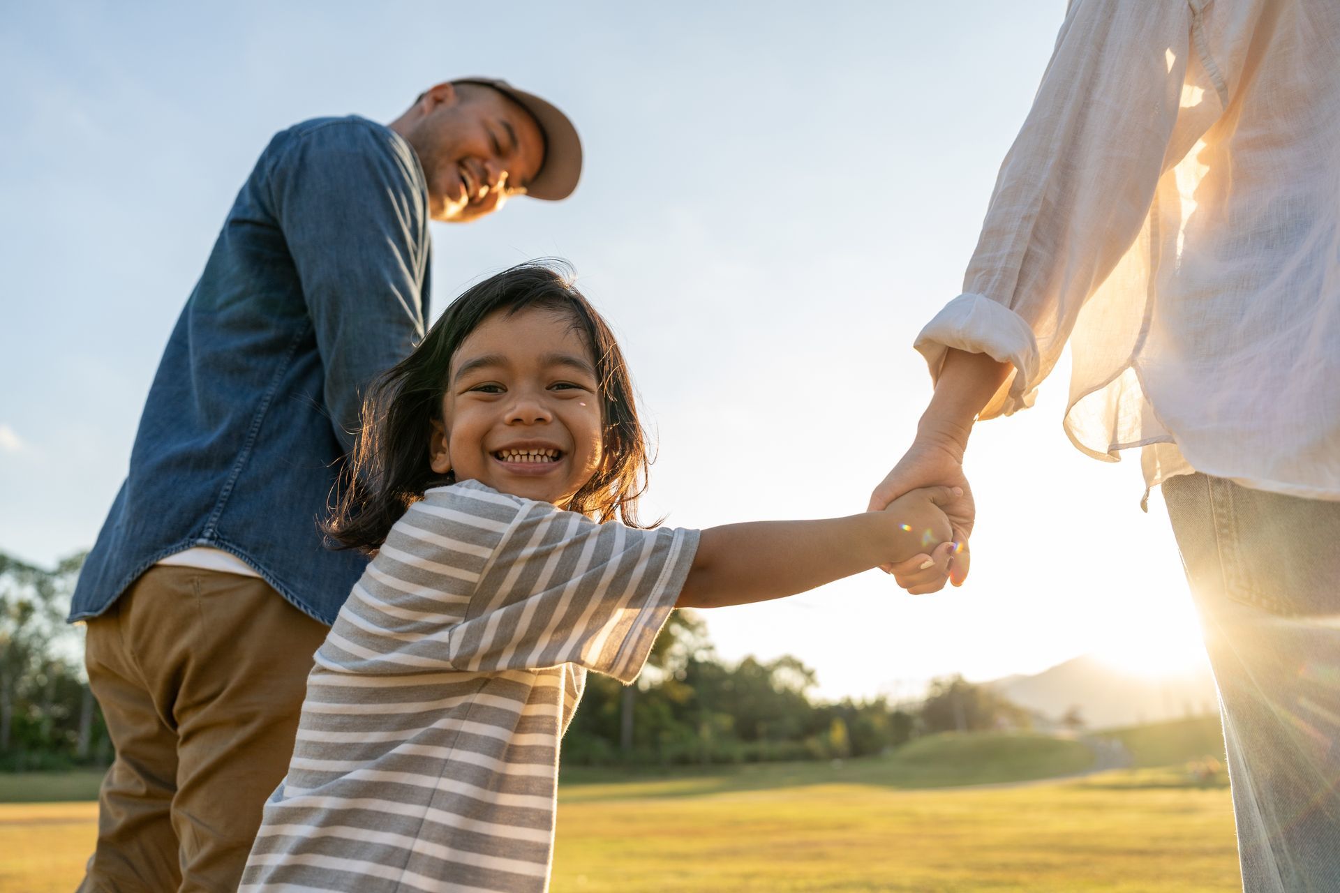 Smiling child holding hands with two adults outdoors at sunset
