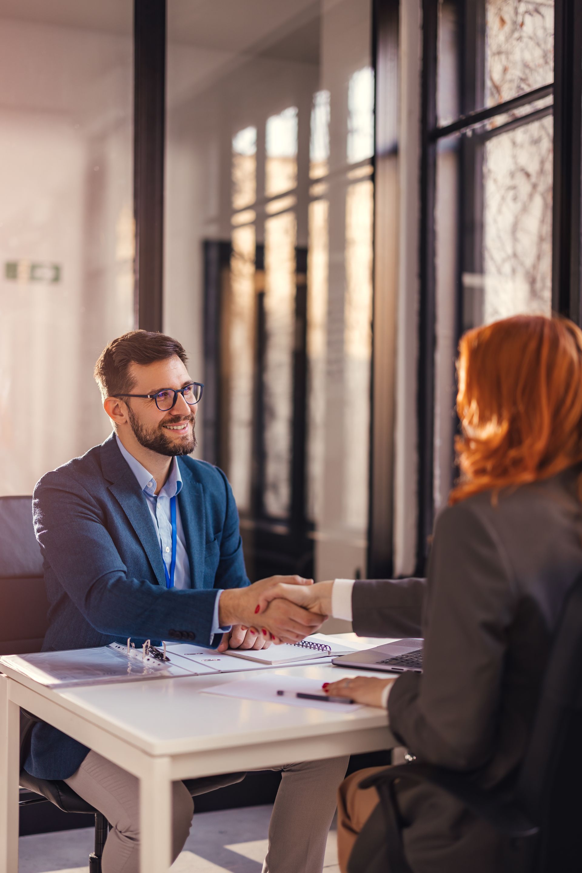 Two people in a bright office shaking hands across a desk, discussing business.