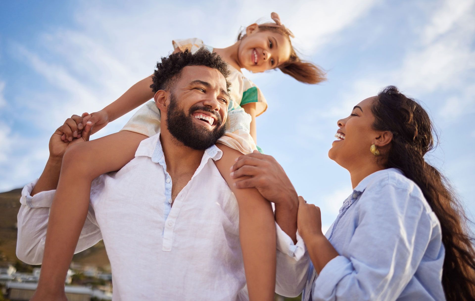 Smiling family outdoors at the beach, with a child on shoulders under a bright sky
