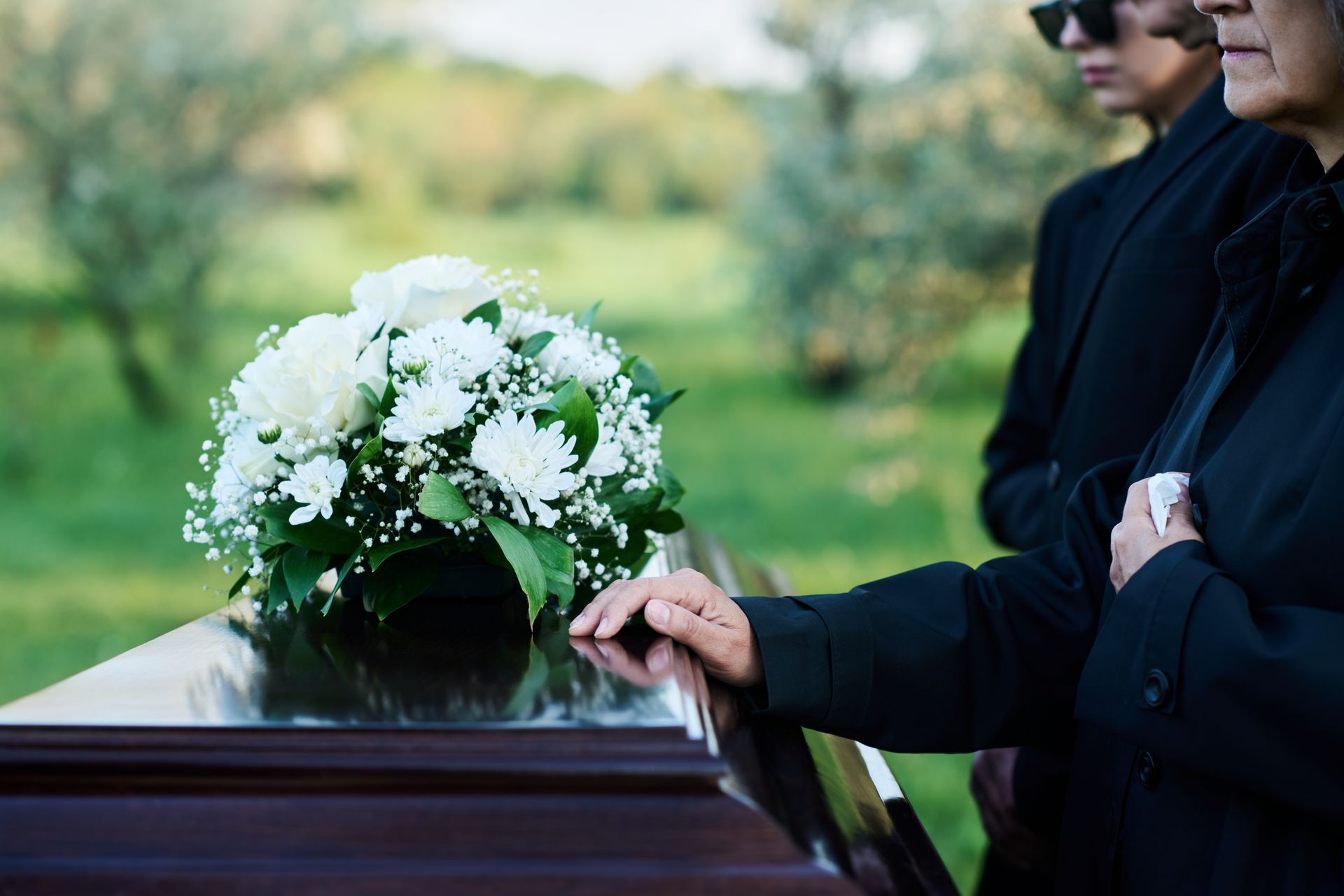 Mourners place a hand on a casket adorned with white flowers in a cemetery